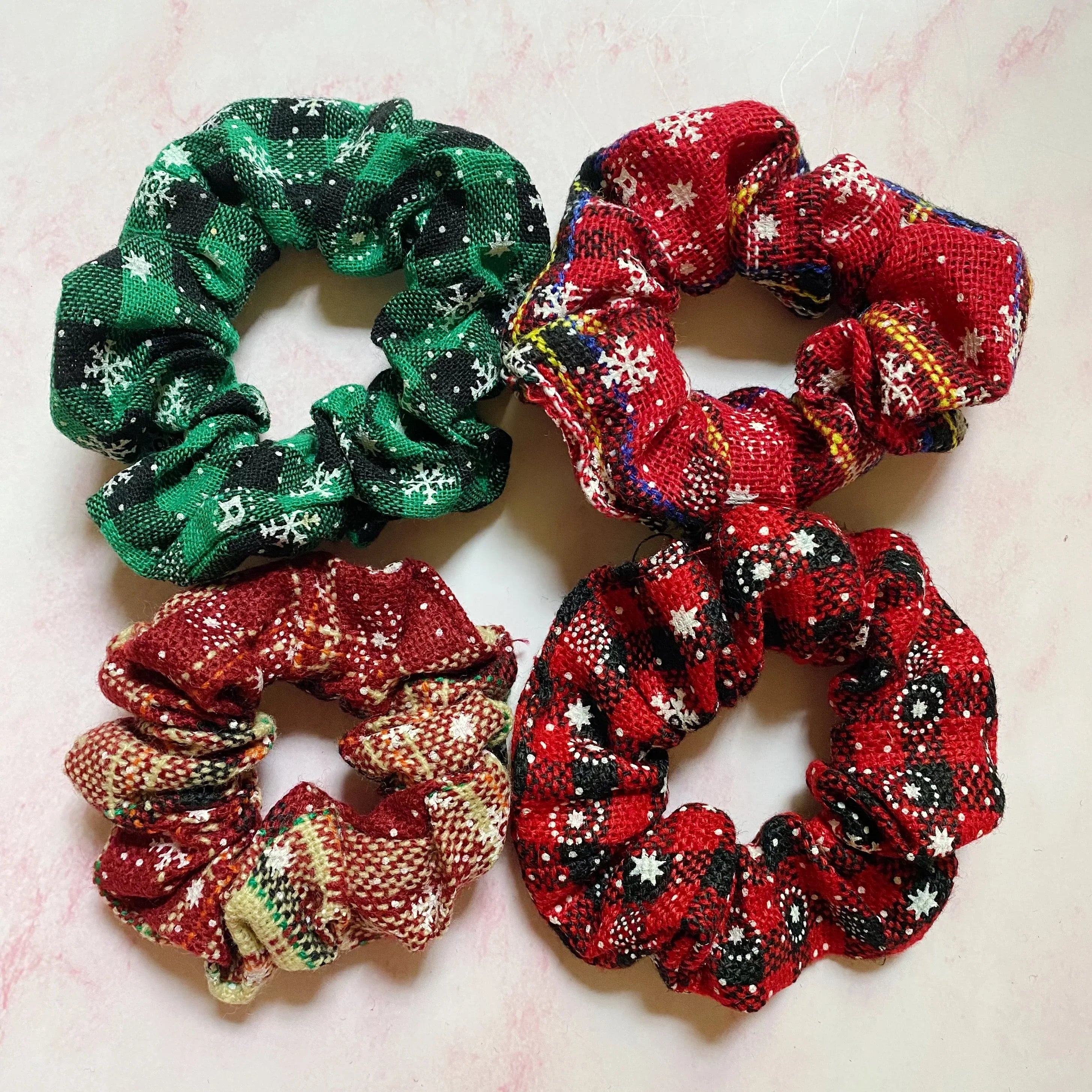 Four festive fabric scrunchies with snowflake patterns in red, green, and plaid, on a pink surface