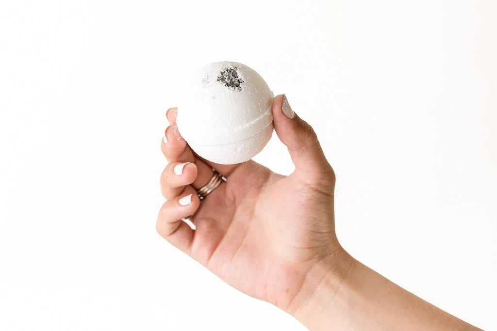Hand holding white round bath bomb with black salt crystals against white background