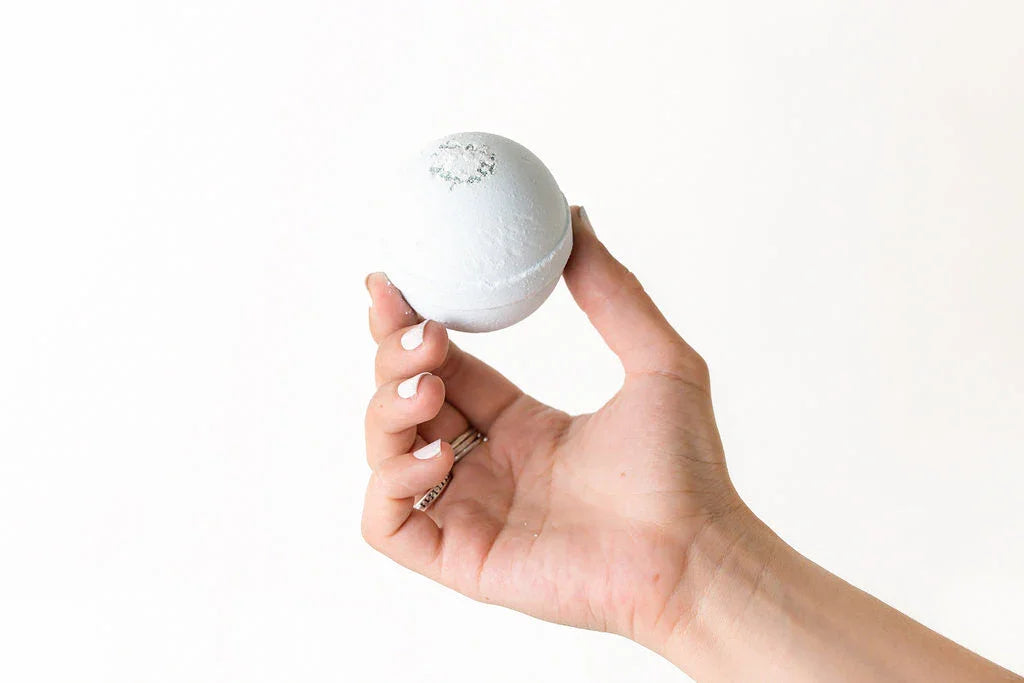 Hand with white nail polish holding a white bath bomb with a silver sparkle detail on a white background
