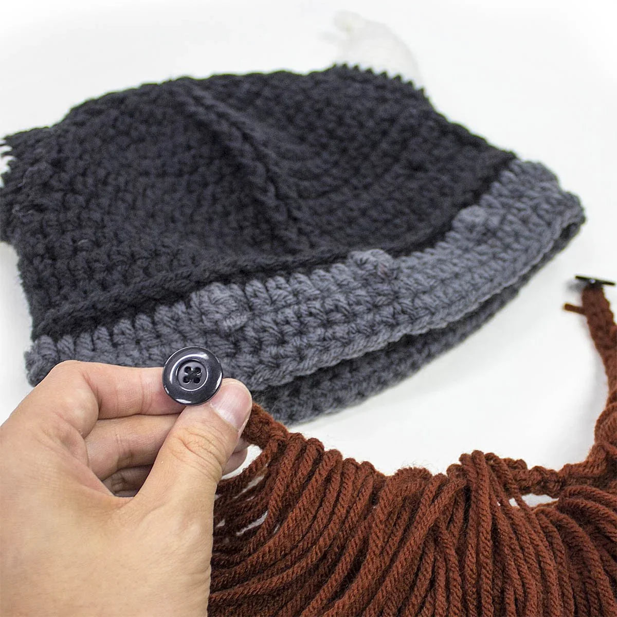 Hand holding button to attach brown yarn beard to black and gray crochet hat