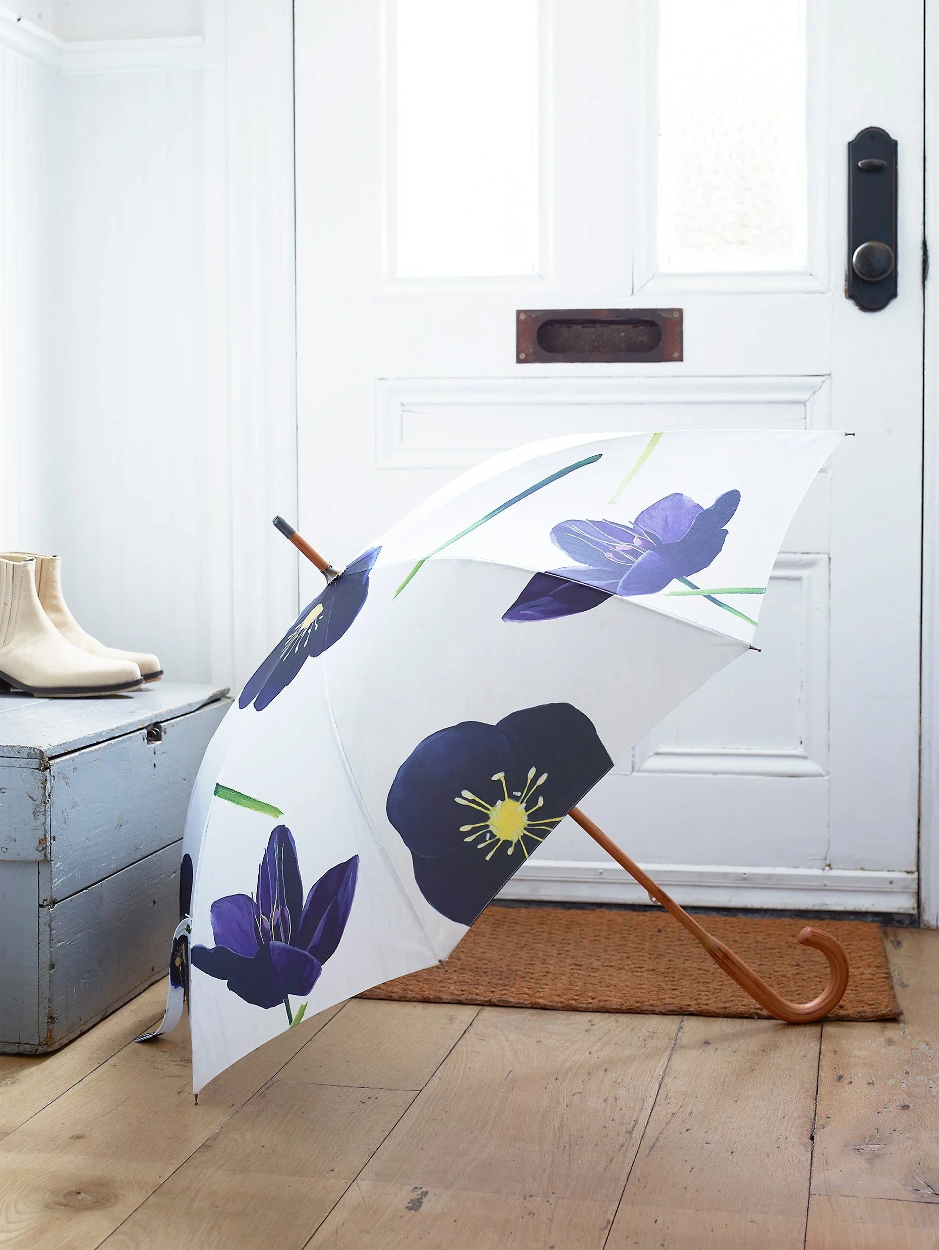 White umbrella with large purple black hellebore flower print, wooden handle on wooden floor by white door
