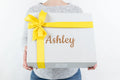 Person holding luxury gift box with yellow ribbon and name Ashley on white patterned wrapping
