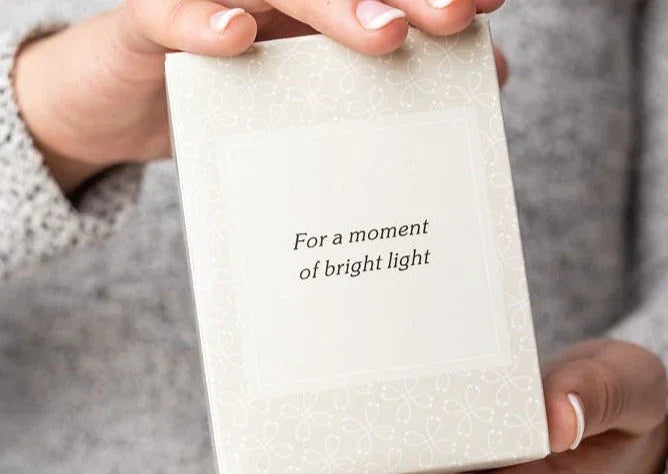 Hand holding a decorative white box with the text 'For a moment of bright light' on the front