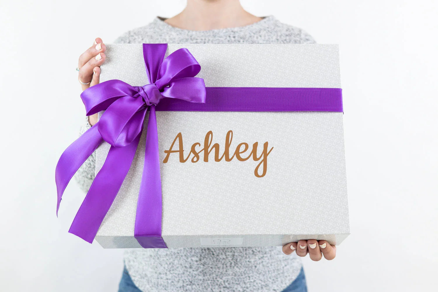 Person holding personalized gift box with purple ribbon and name Ashley on gray patterned wrapping