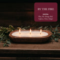 Rustic wooden three-wick candle by Qu!rk Boutique, lit on a wooden table with greenery, 'By The Fire' scent.