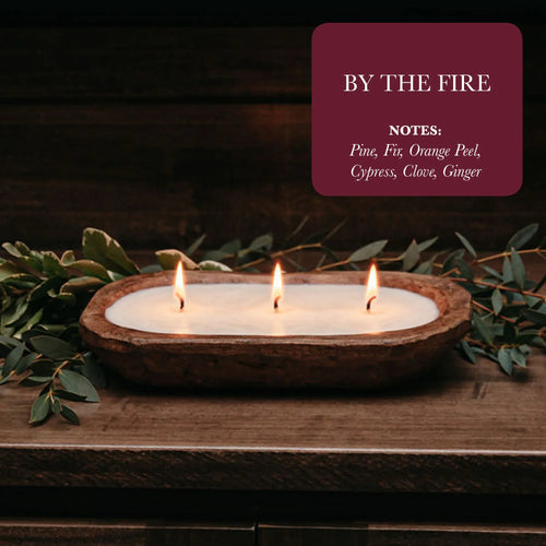 Rustic wooden three-wick candle by Qu!rk Boutique, lit on a wooden table with greenery, 'By The Fire' scent.