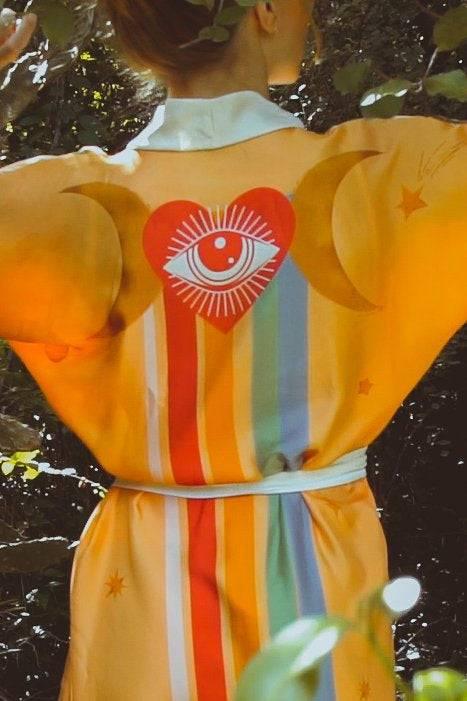 Person wearing a yellow robe with colorful stripes, a red heart with an eye, crescent moons, and stars in a leafy outdoor setting