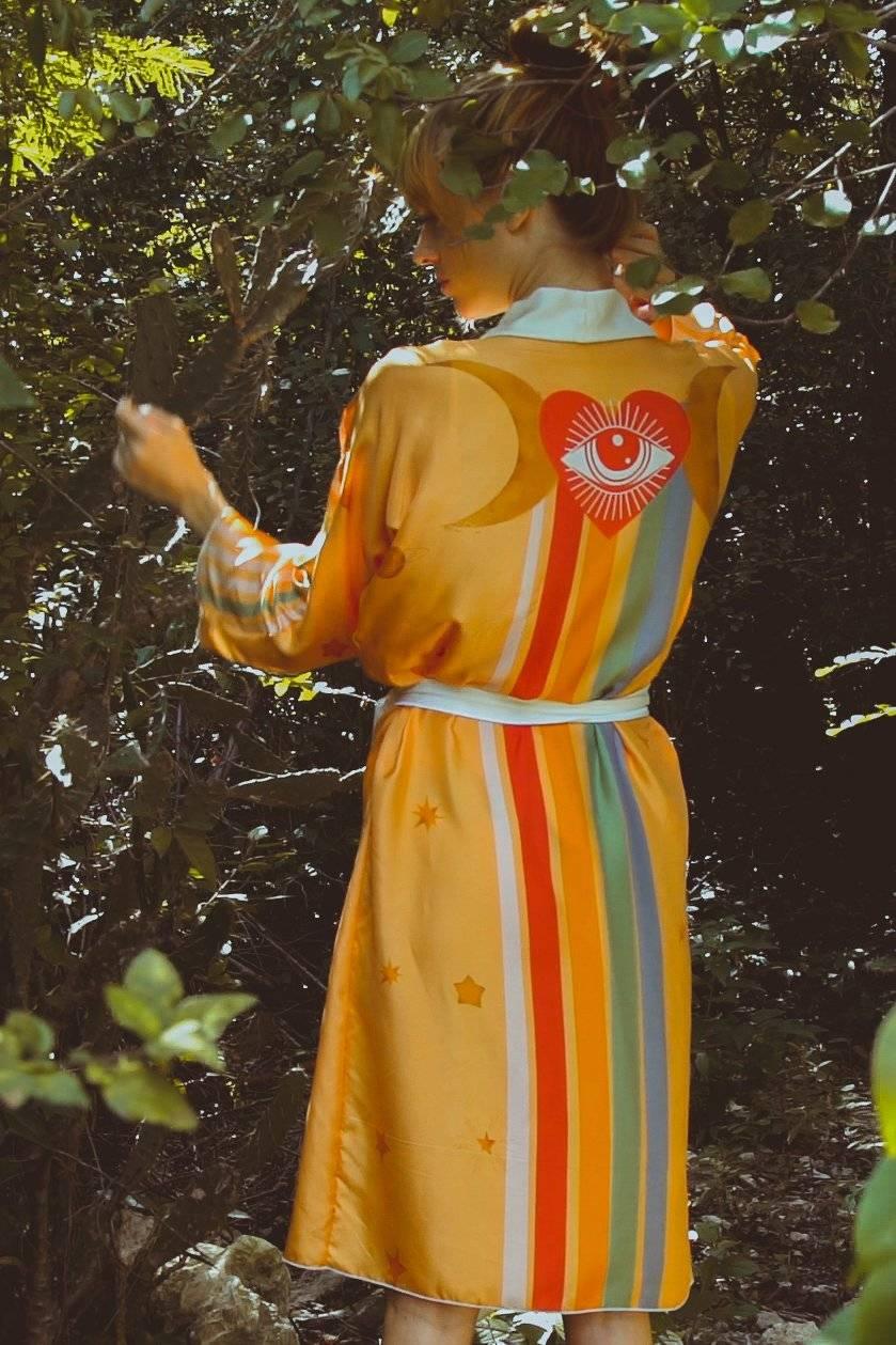 Woman in yellow robe with eye and heart graphic, colorful stripes, and star patterns in forest