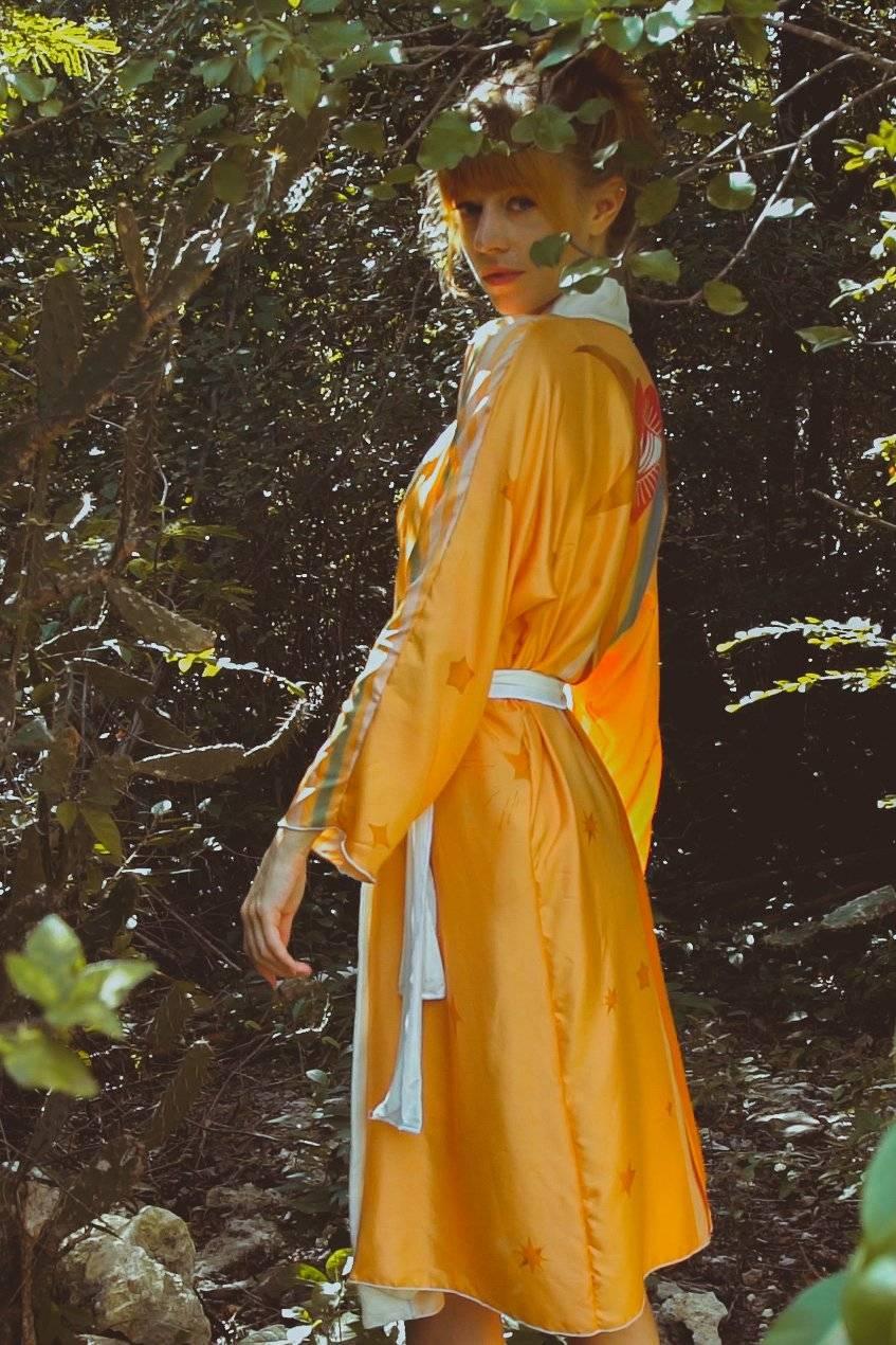 Woman wearing an orange kimono with star patterns and white belt standing in a forest setting
