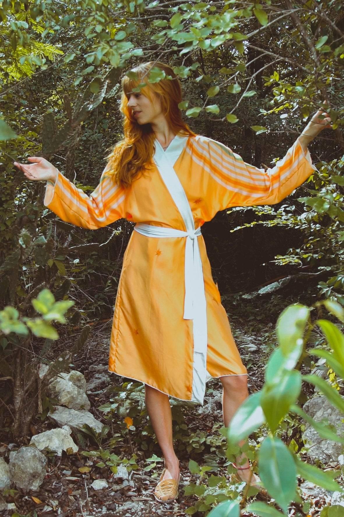 Woman in orange and white kimono-style robe standing amidst green foliage in a forest setting