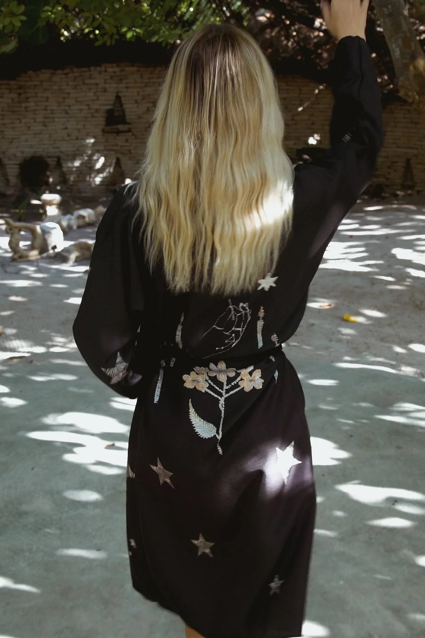 Blonde woman in black dress with floral and star embroidery standing outdoors under tree shadows