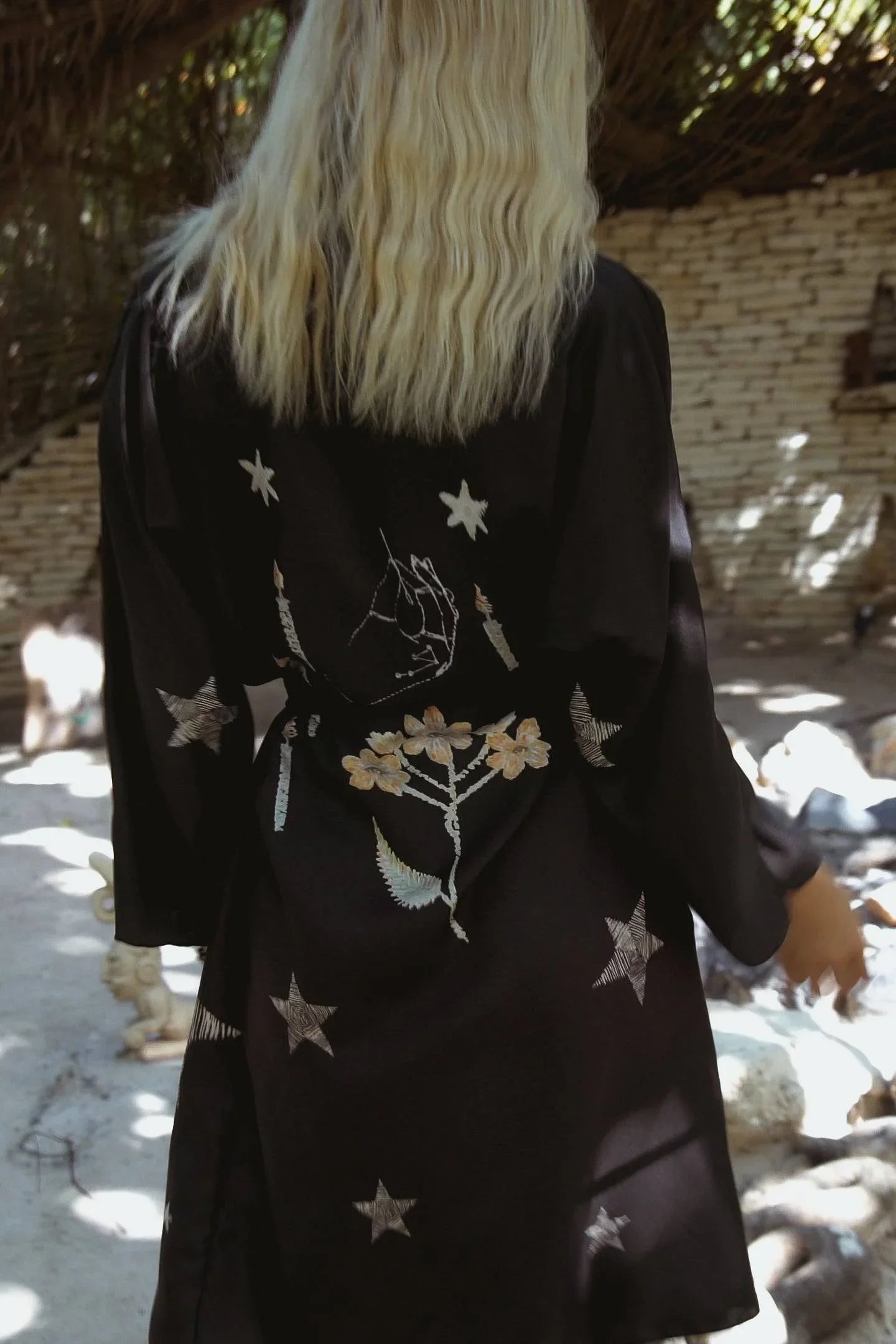 Blonde woman wearing a black robe with star, candle, flower, and hand embroidery in outdoor setting
