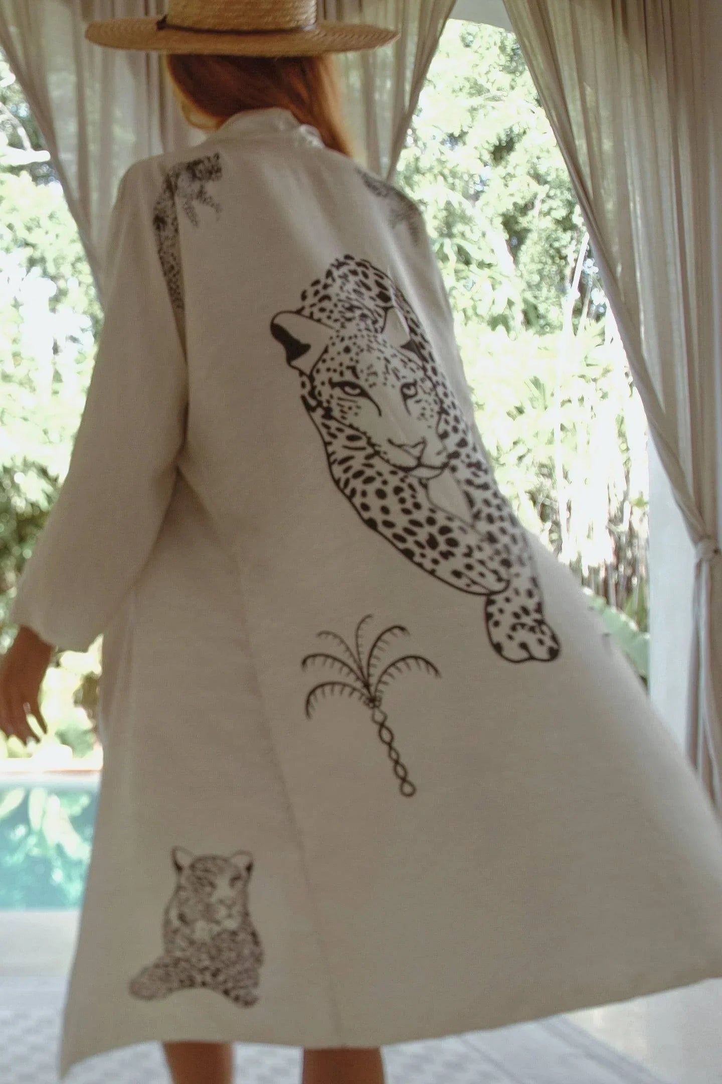 Woman wearing a white robe with black leopard and palm tree prints, standing by a pool