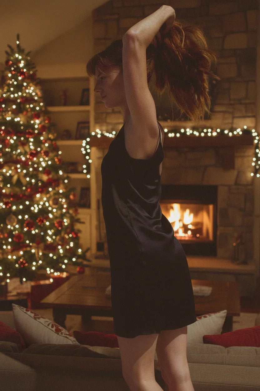 Woman in black dress tying hair by decorated Christmas tree and lit fireplace at home