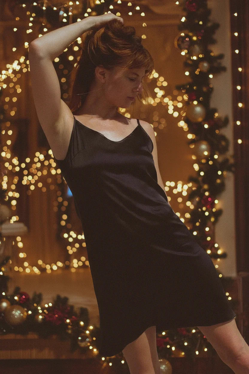 Woman in black slip dress posing indoors with festive Christmas lights and decorated tree background