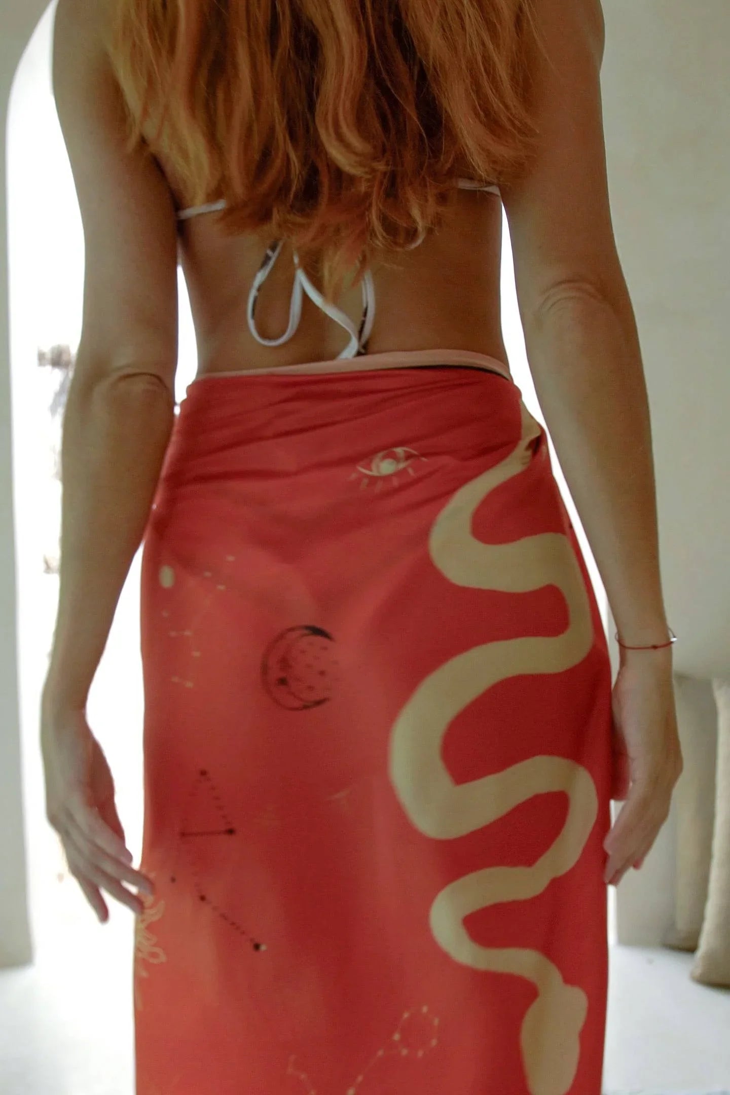 Woman with long hair wearing a red sarong with eye and snake designs, standing indoors