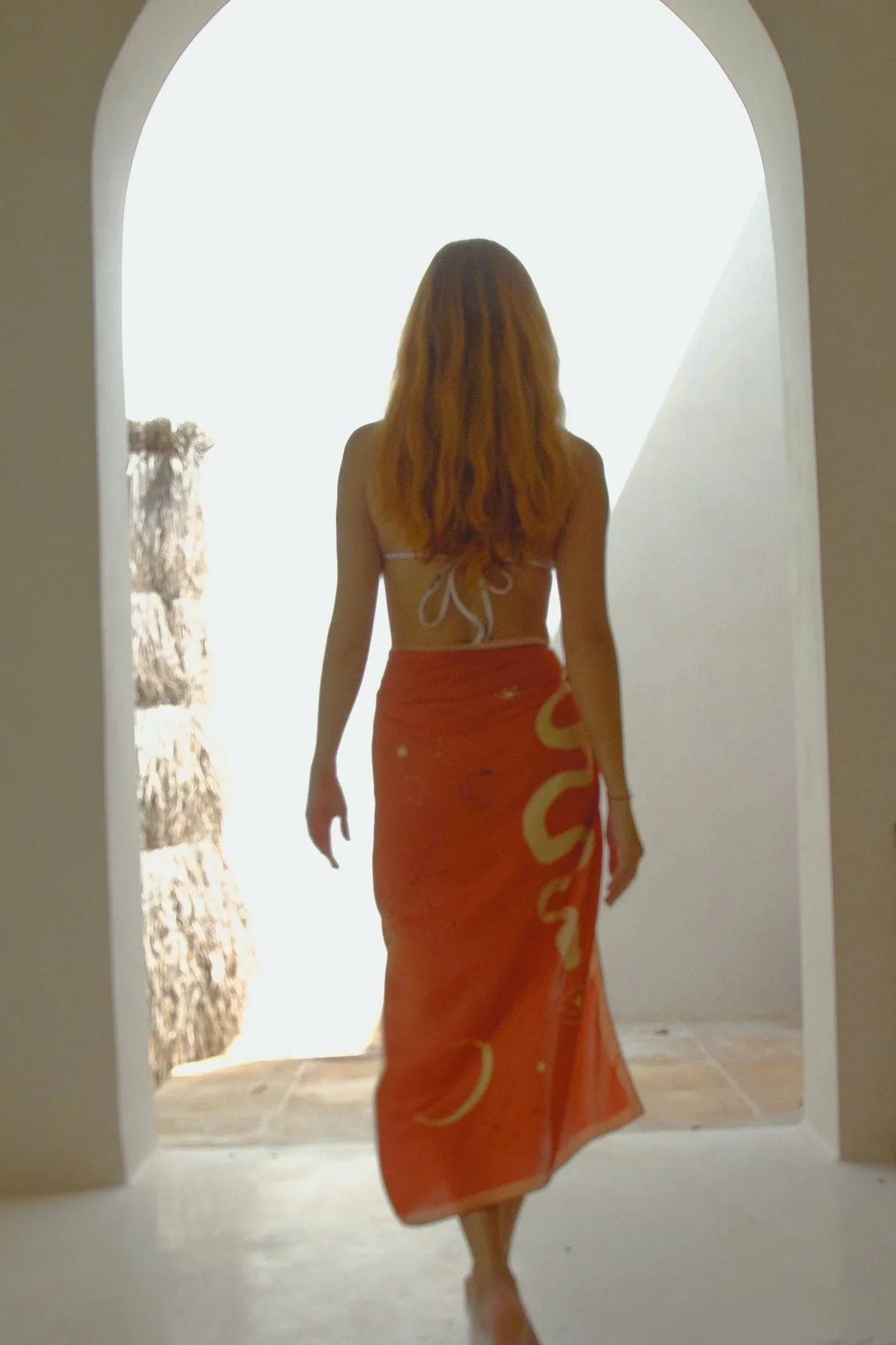 Woman with long hair wearing a white bikini top and orange sarong standing in arched doorway with bright light