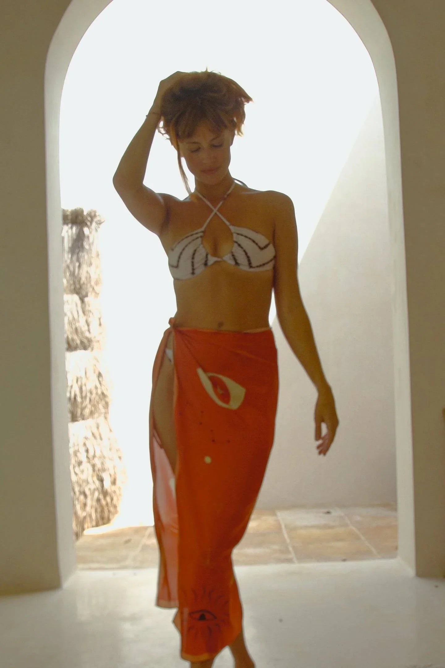 Woman posing in sunlight wearing a white halter bikini top and an orange sarong with eye designs