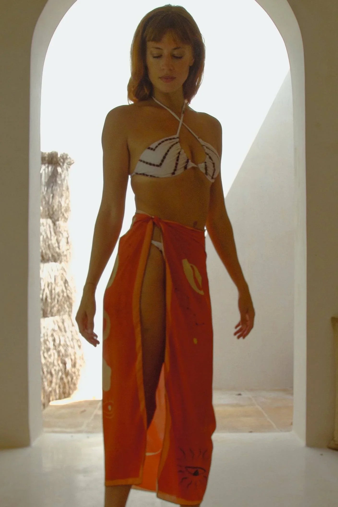 Woman wearing white patterned bikini top and orange sarong in sunlit archway