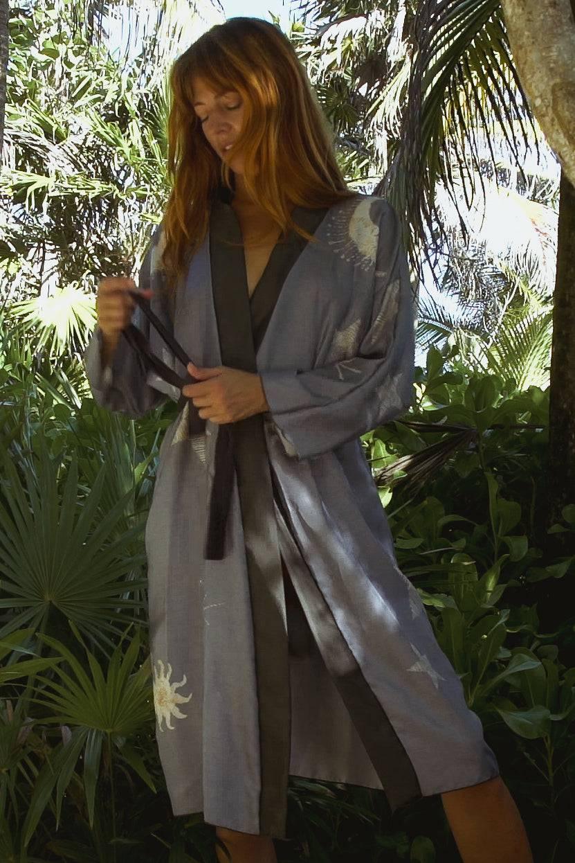 Woman in a gray sun-patterned robe tying the belt in a lush tropical garden setting