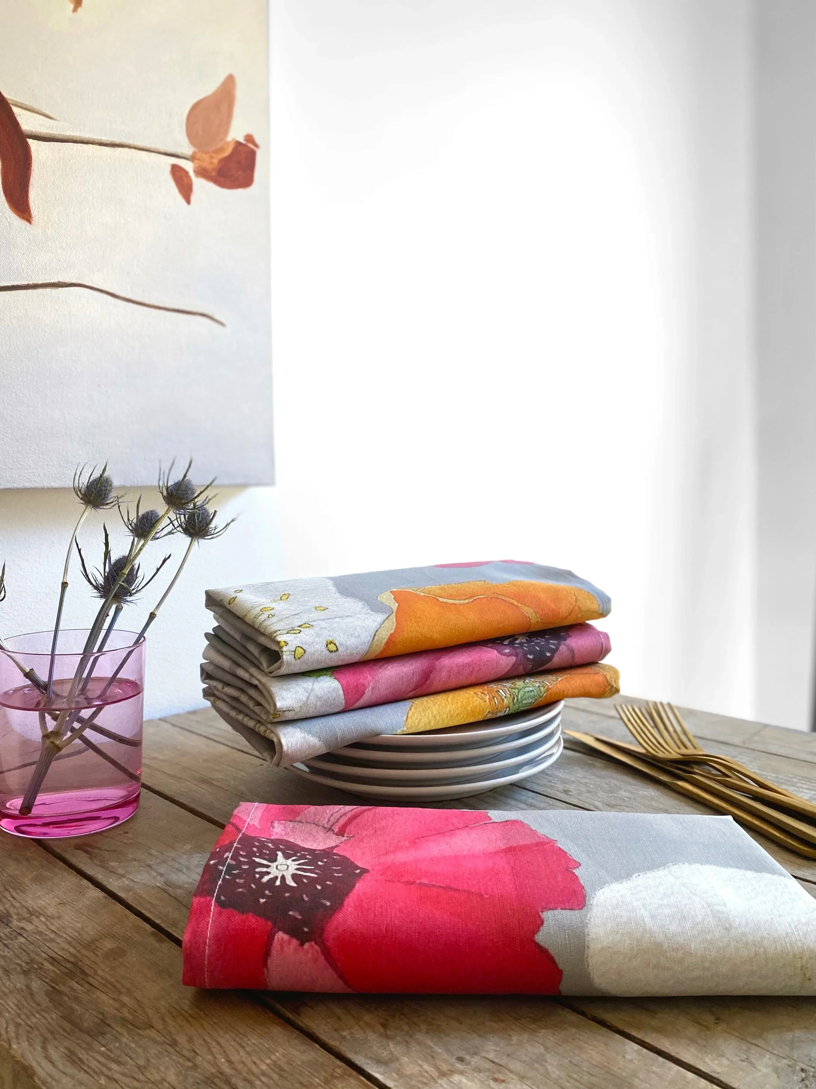 Stack of colorful floral napkins on white plates with gold forks and a pink vase of dried flowers on wooden table