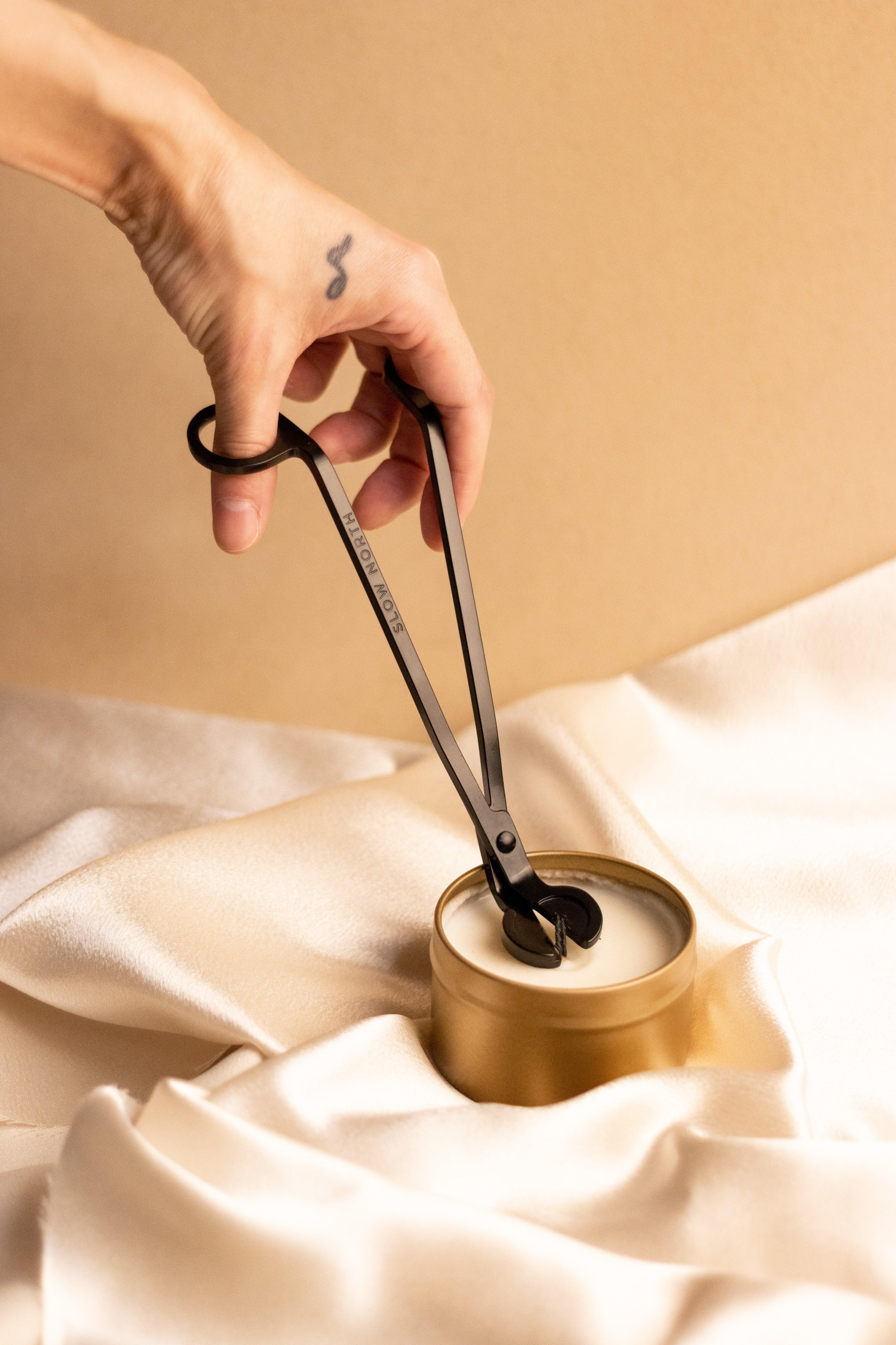 Hand holding black wick trimmer above gold candle on cream satin fabric backdrop