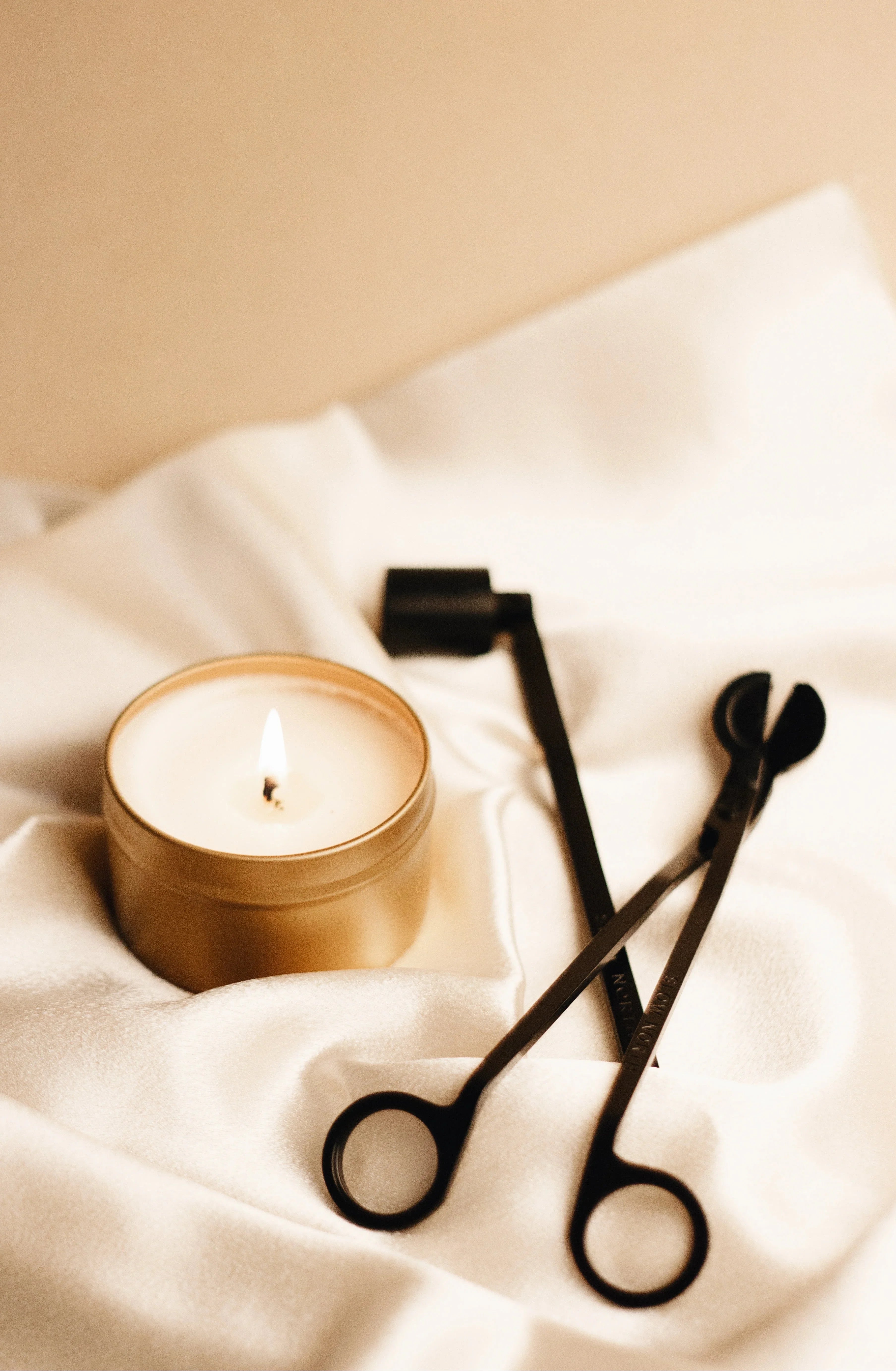 Lit candle in a gold container with black wick trimmer and snuffer on white fabric