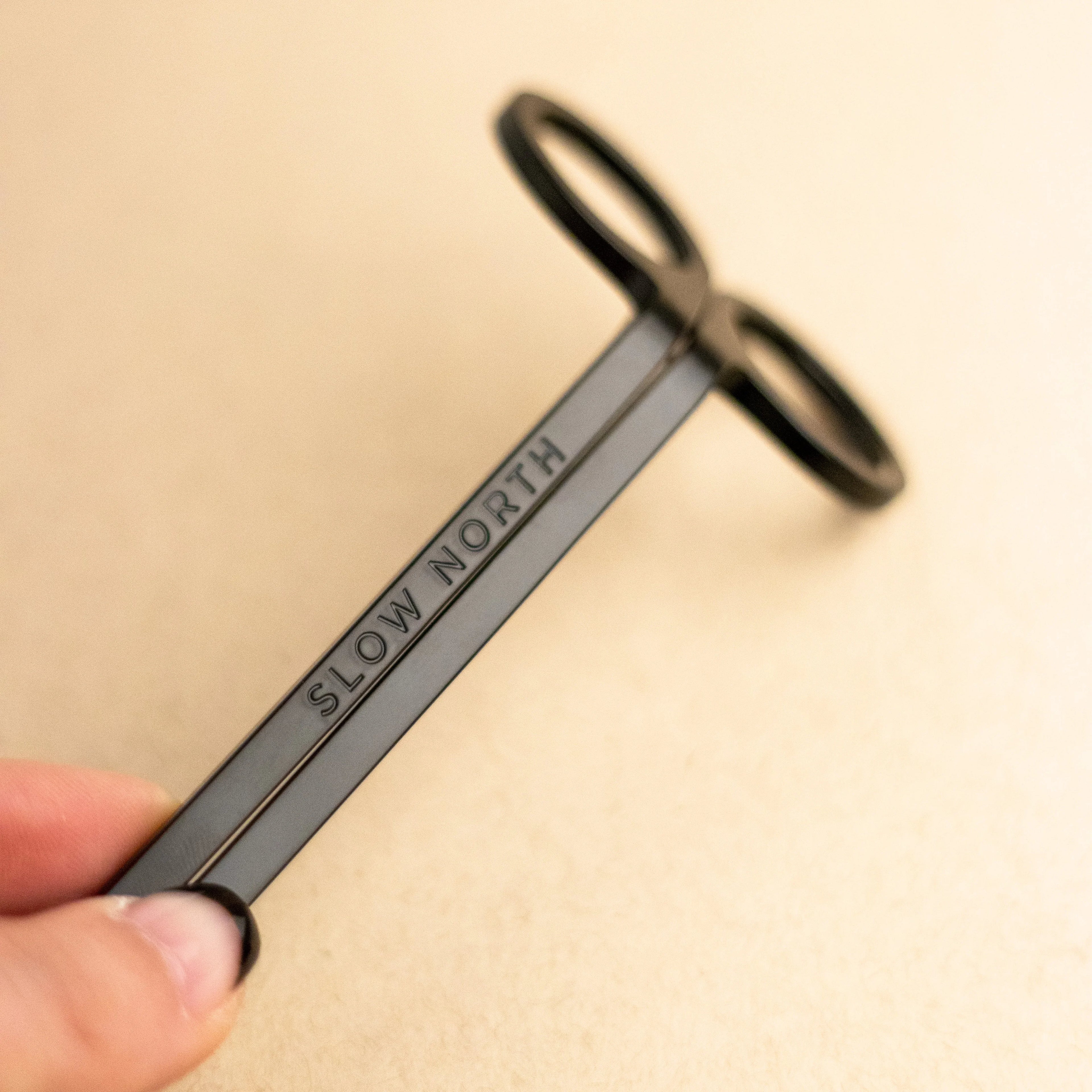Hand holding black metal wick trimmer engraved with Slow North on a beige background