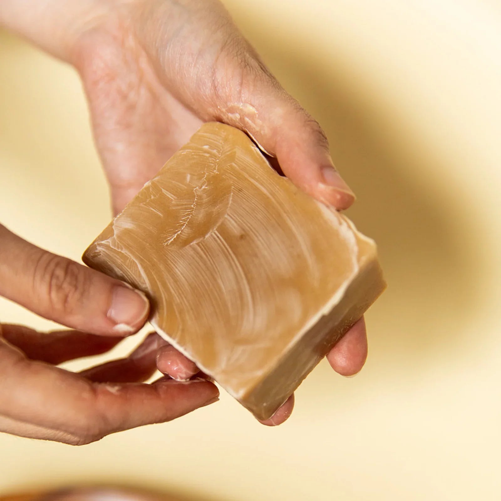 Hands holding a square coconut oatmeal natural soap bar with creamy texture on beige background