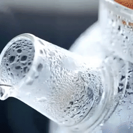 Close-up of a glass teapot pouring hot water, with steam and droplets at Qu!rk Boutique.