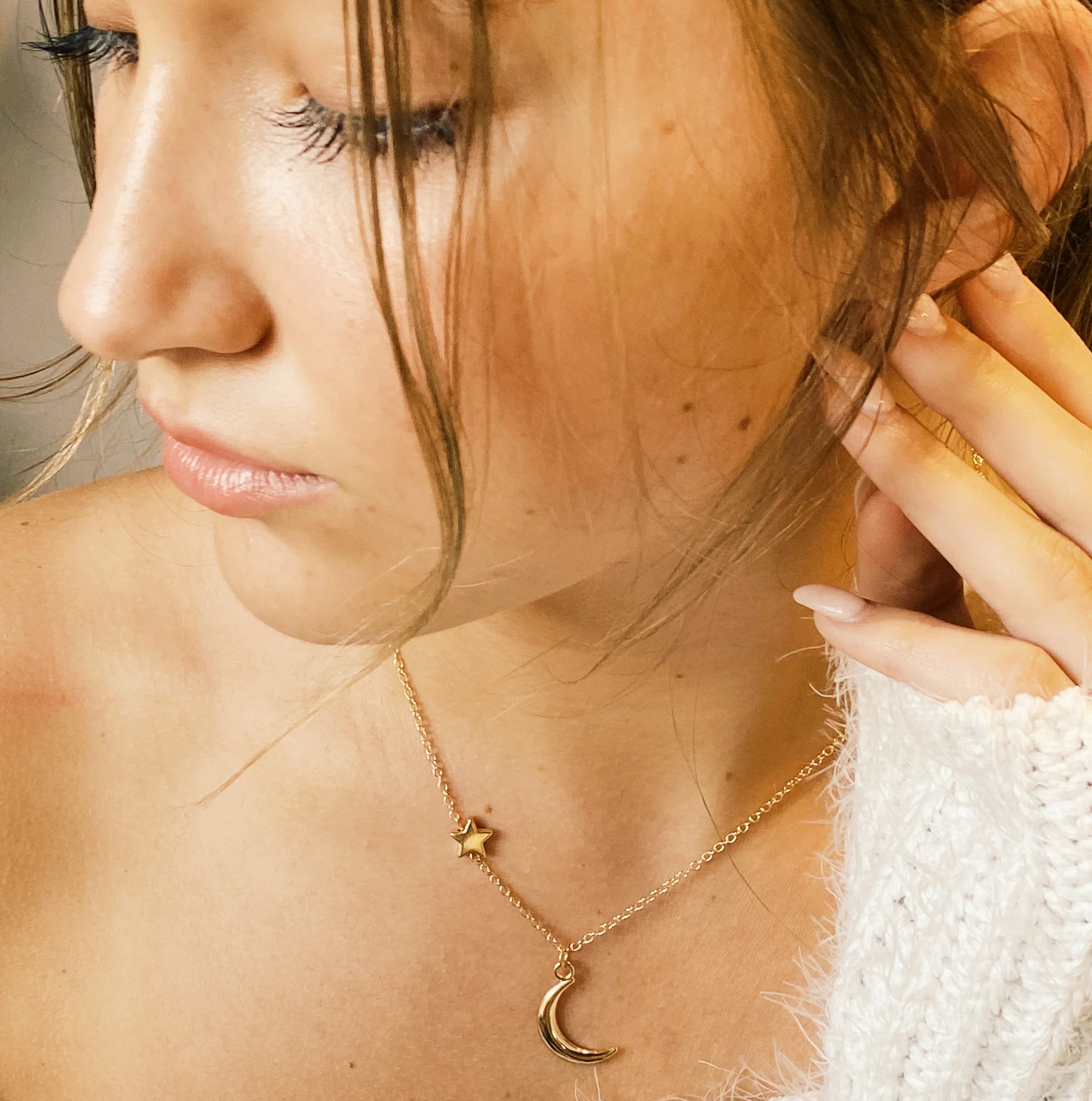Close-up of woman wearing gold star and crescent moon necklace with soft natural makeup and cozy white sweater