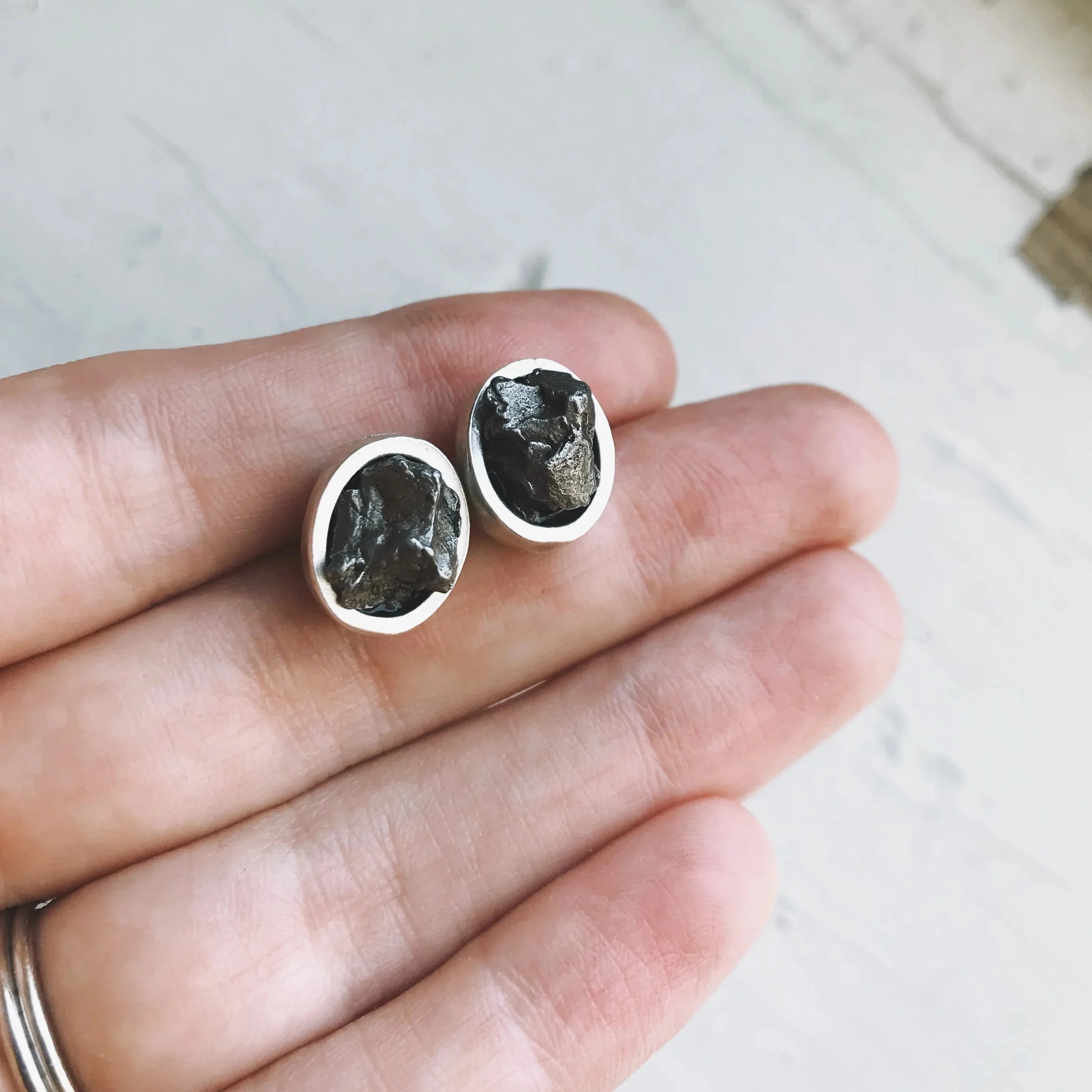 Hand holding two rustic raw black stone stud earrings with silver oval settings on white background