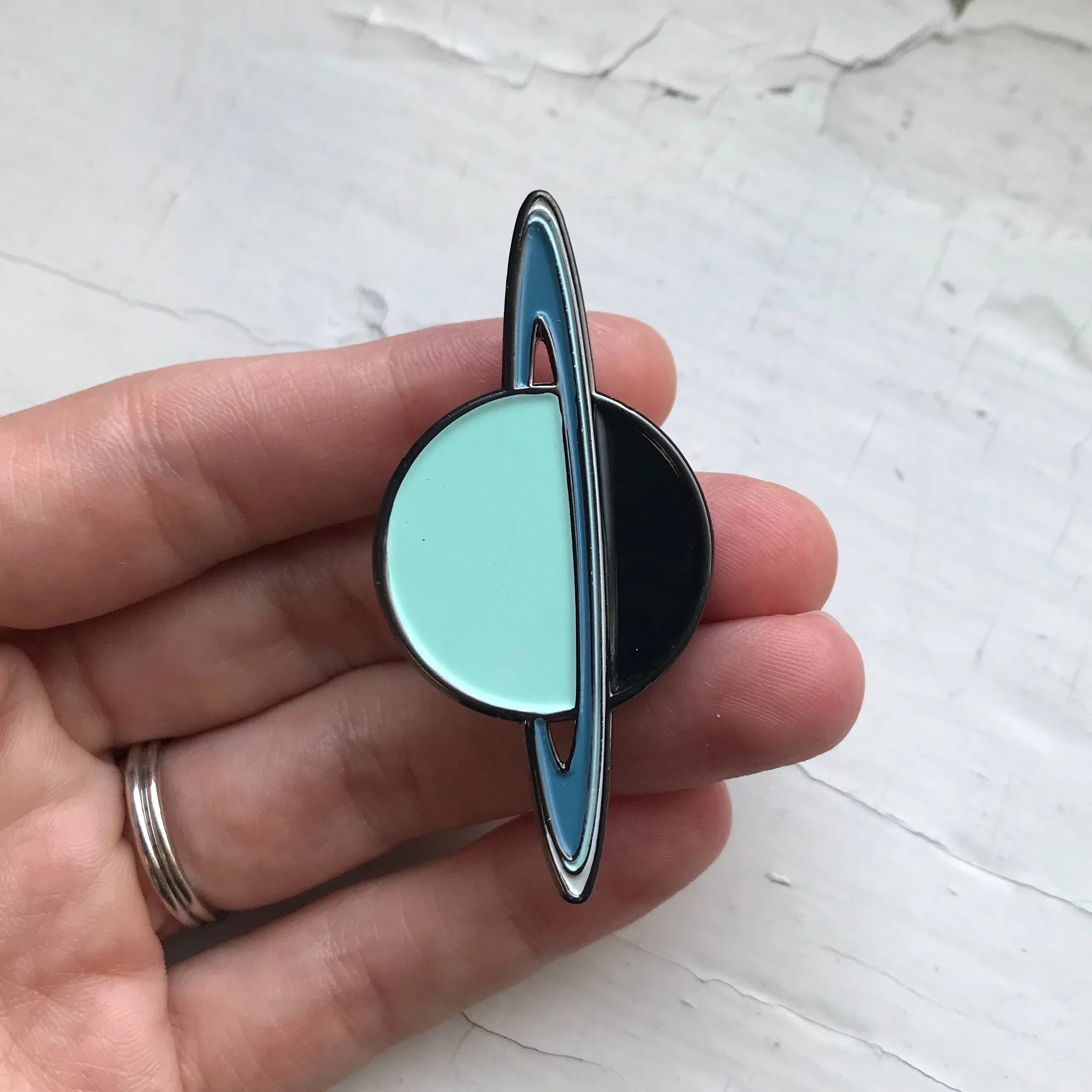 Hand holding enamel pin of a stylized planet with blue rings and half turquoise, half black circle