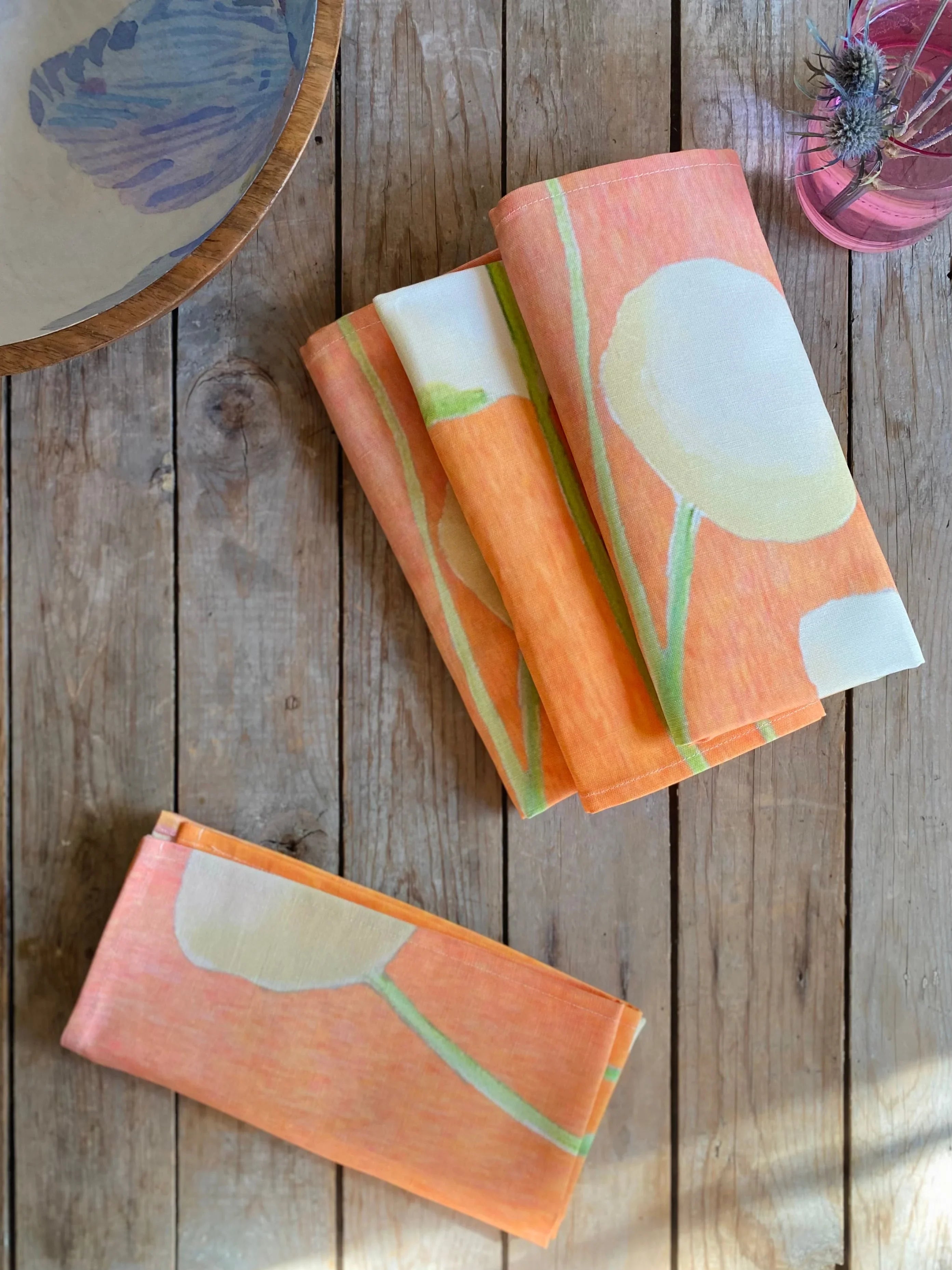 Folded orange floral print linen napkins on rustic wooden table with ceramic dish and pink vase