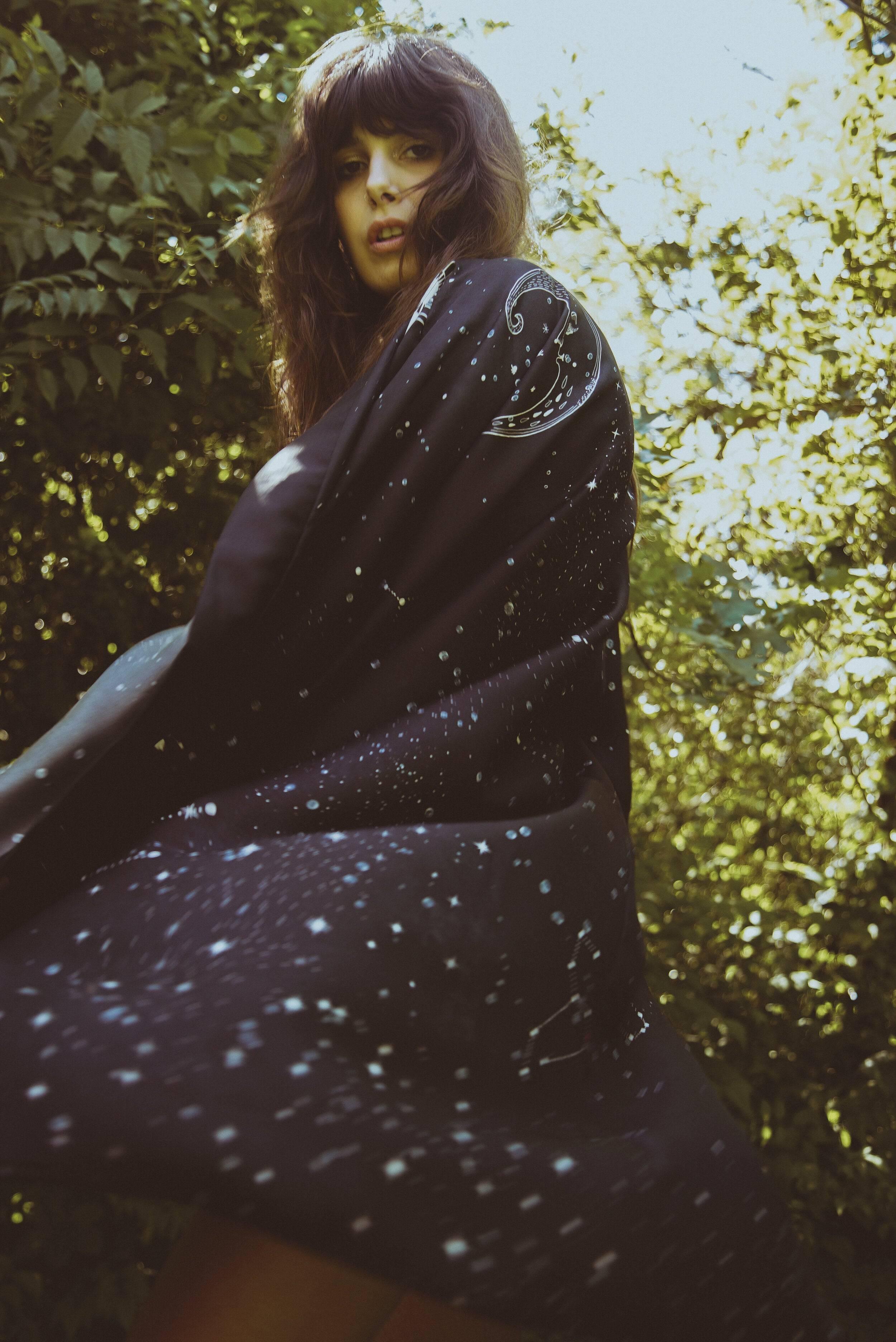 Woman outdoors wearing a black galaxy print shawl with moon and star designs against green foliage