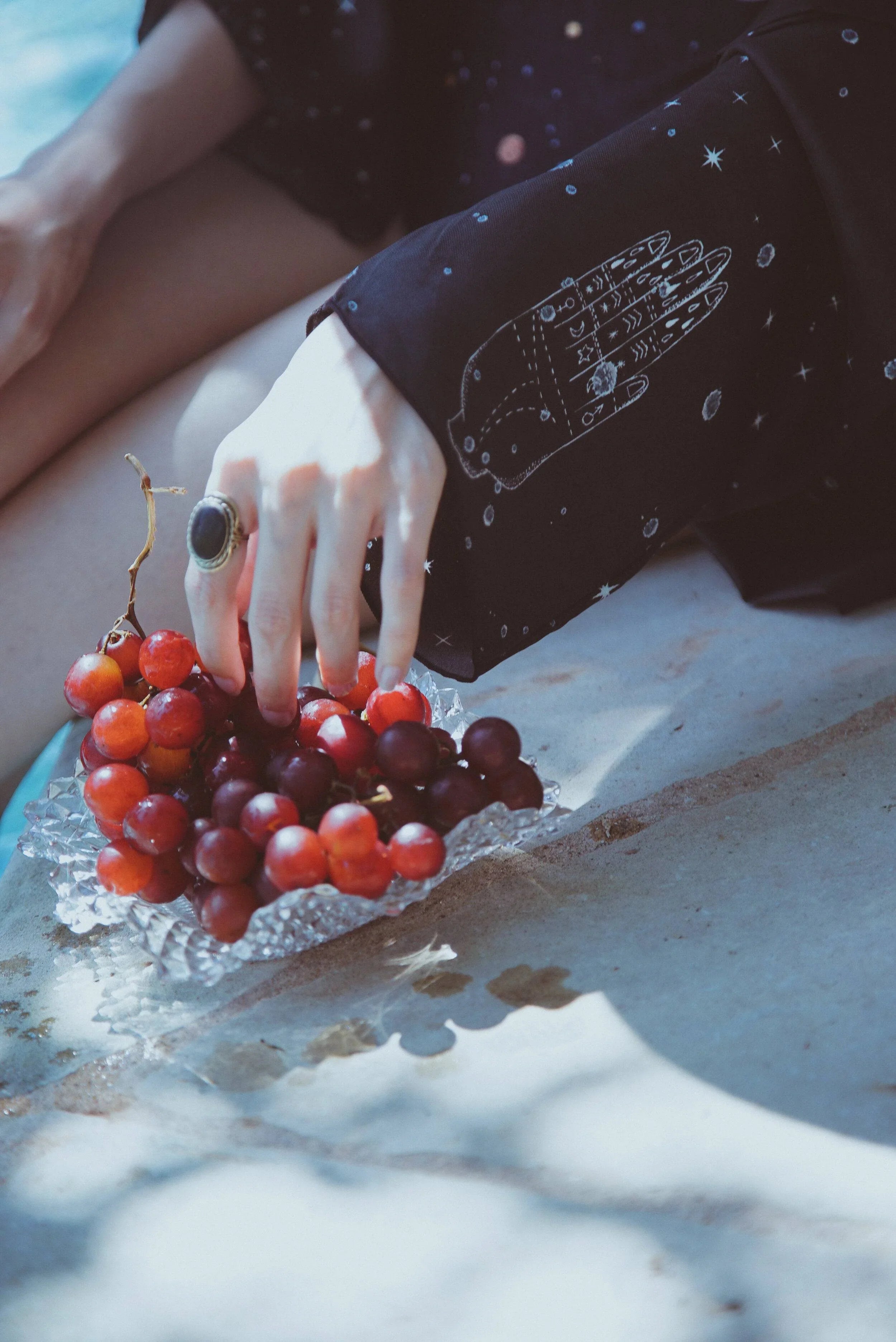 Hand with ring reaching for red grapes on a textured glass dish, black sleeve with star and hand print design
