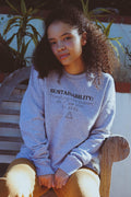 Young woman with curly hair wearing grey sustainability sweatshirt sitting on wooden bench outdoors