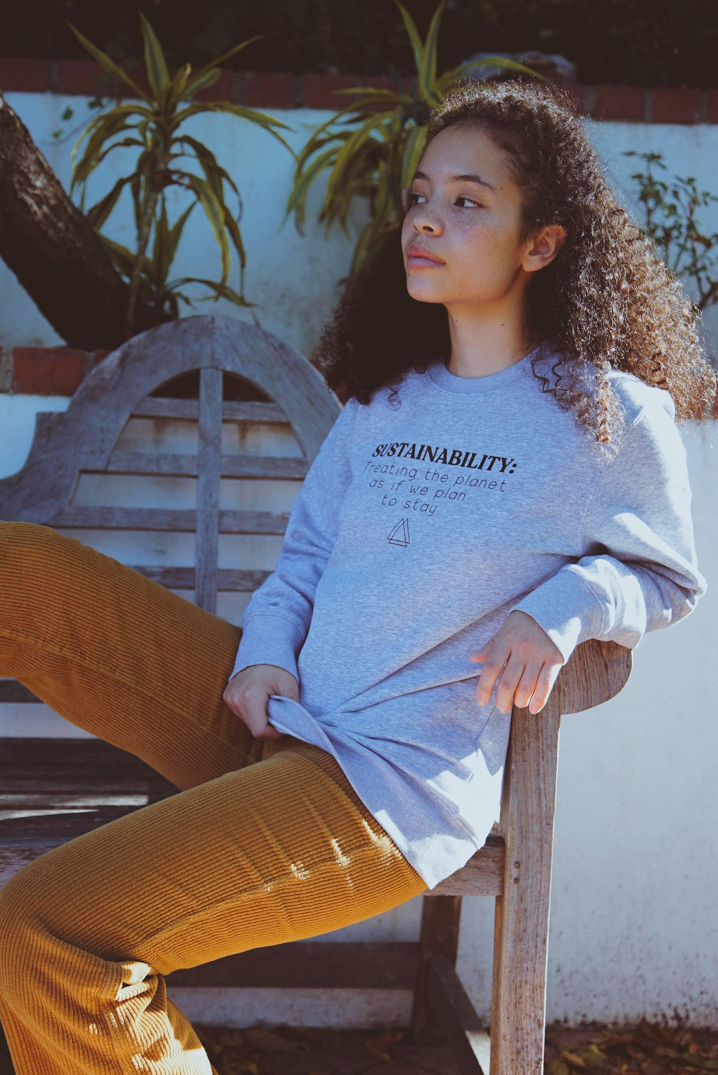 Young woman with curly hair wearing a gray sustainability sweatshirt and mustard pants sitting on wooden bench outdoors