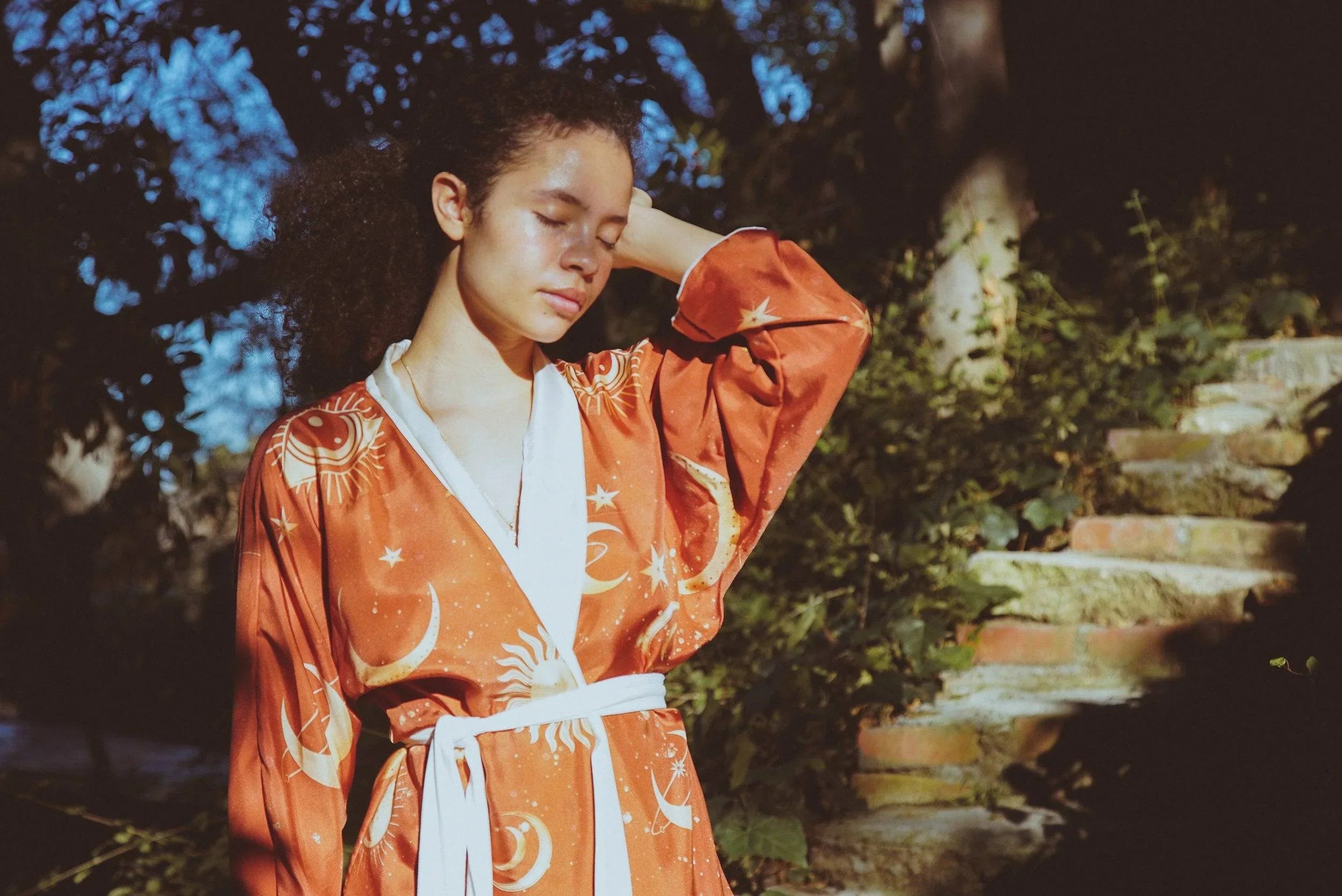 Woman in orange celestial robe standing outdoors near stone steps with eyes closed in sunlight