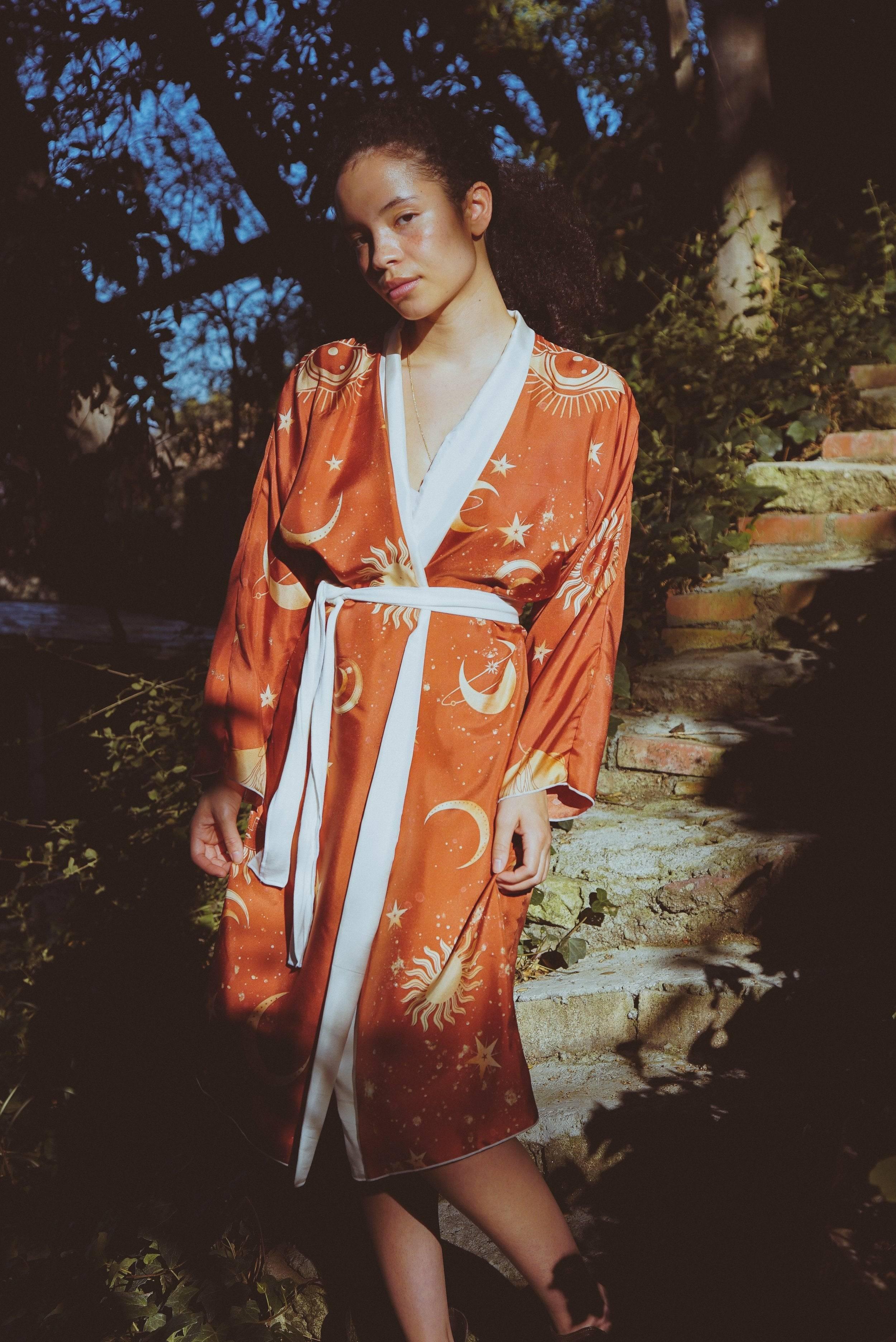 Woman standing outdoors on stone steps wearing an orange robe with sun, moon, and star celestial prints
