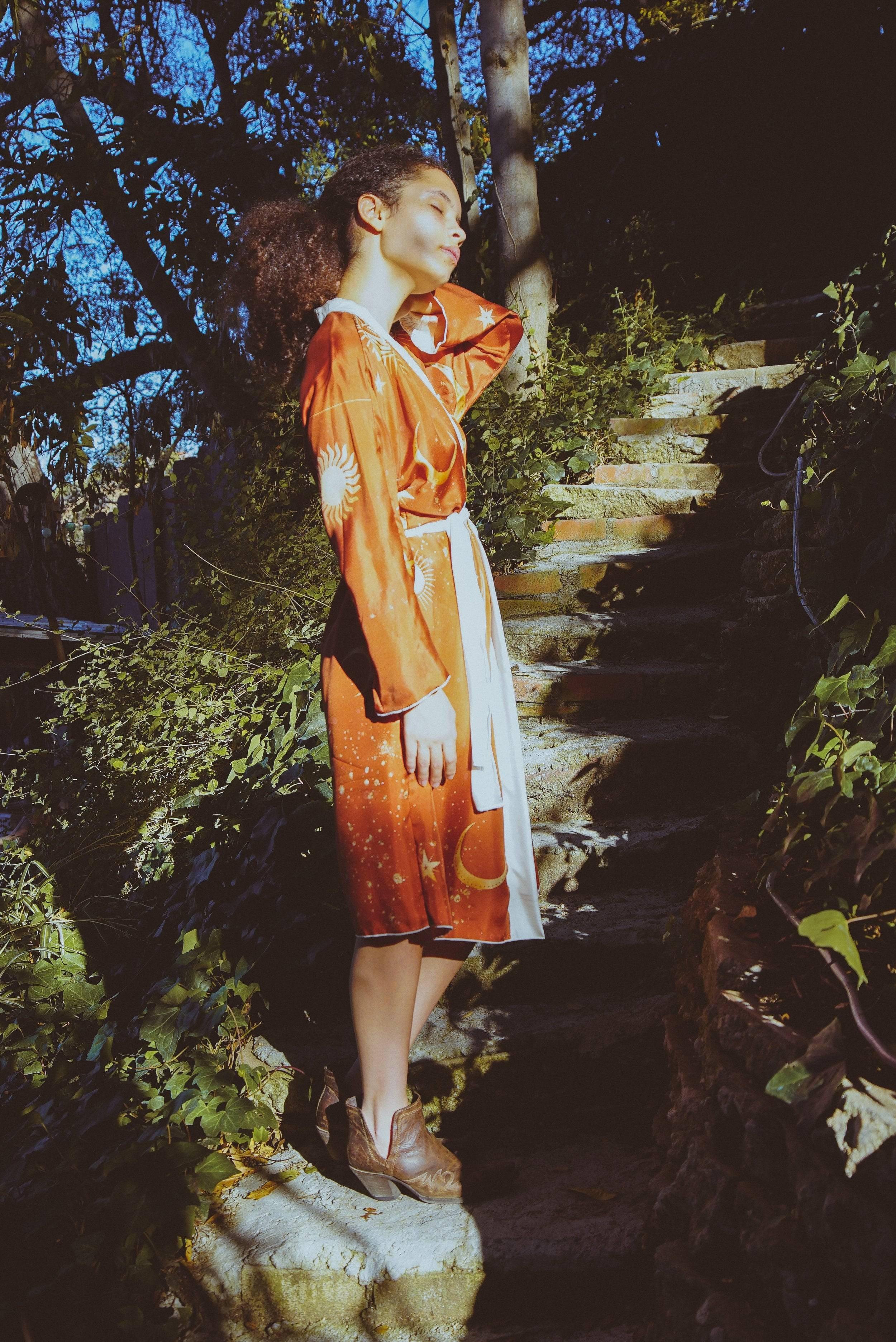 Woman in orange celestial robe and brown boots standing on stone steps surrounded by greenery in sunlight