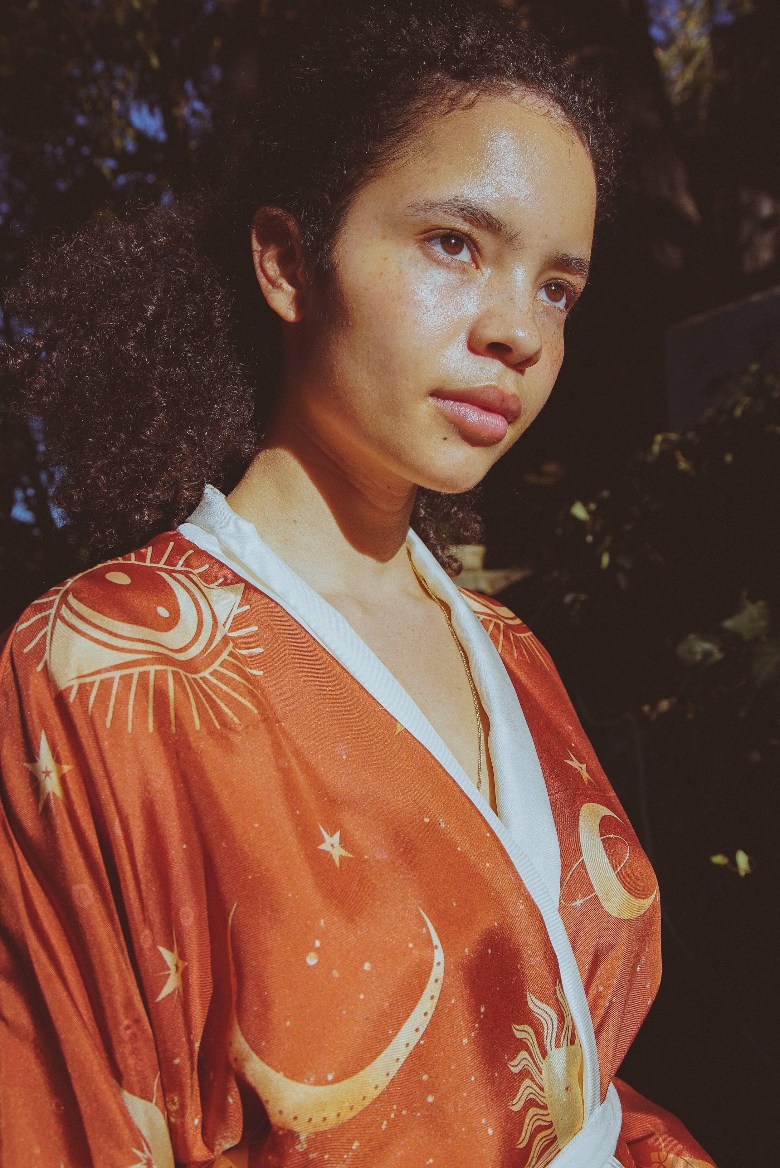 Young woman with curly hair wearing rust-orange celestial robe with sun and moon patterns in natural light