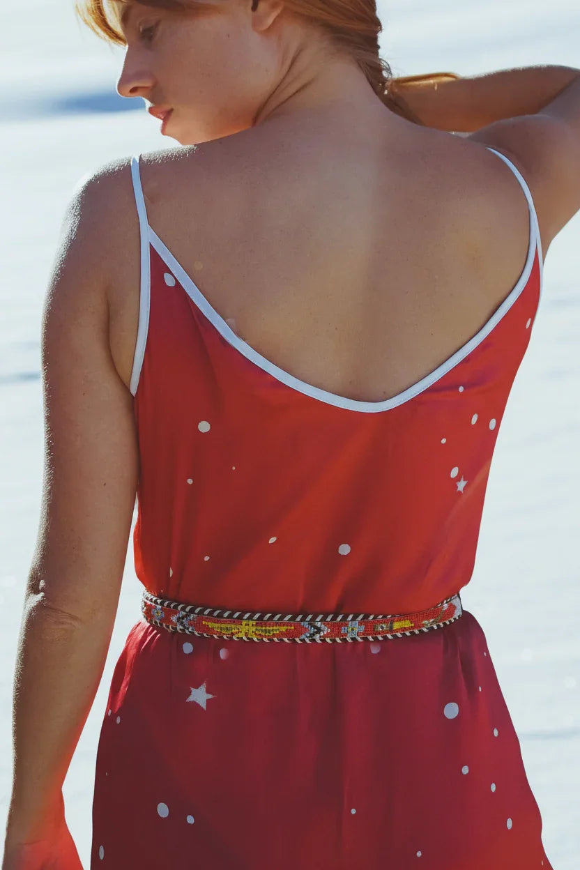 Woman wearing a red star-patterned spaghetti strap dress with a colorful beaded belt, standing by the water