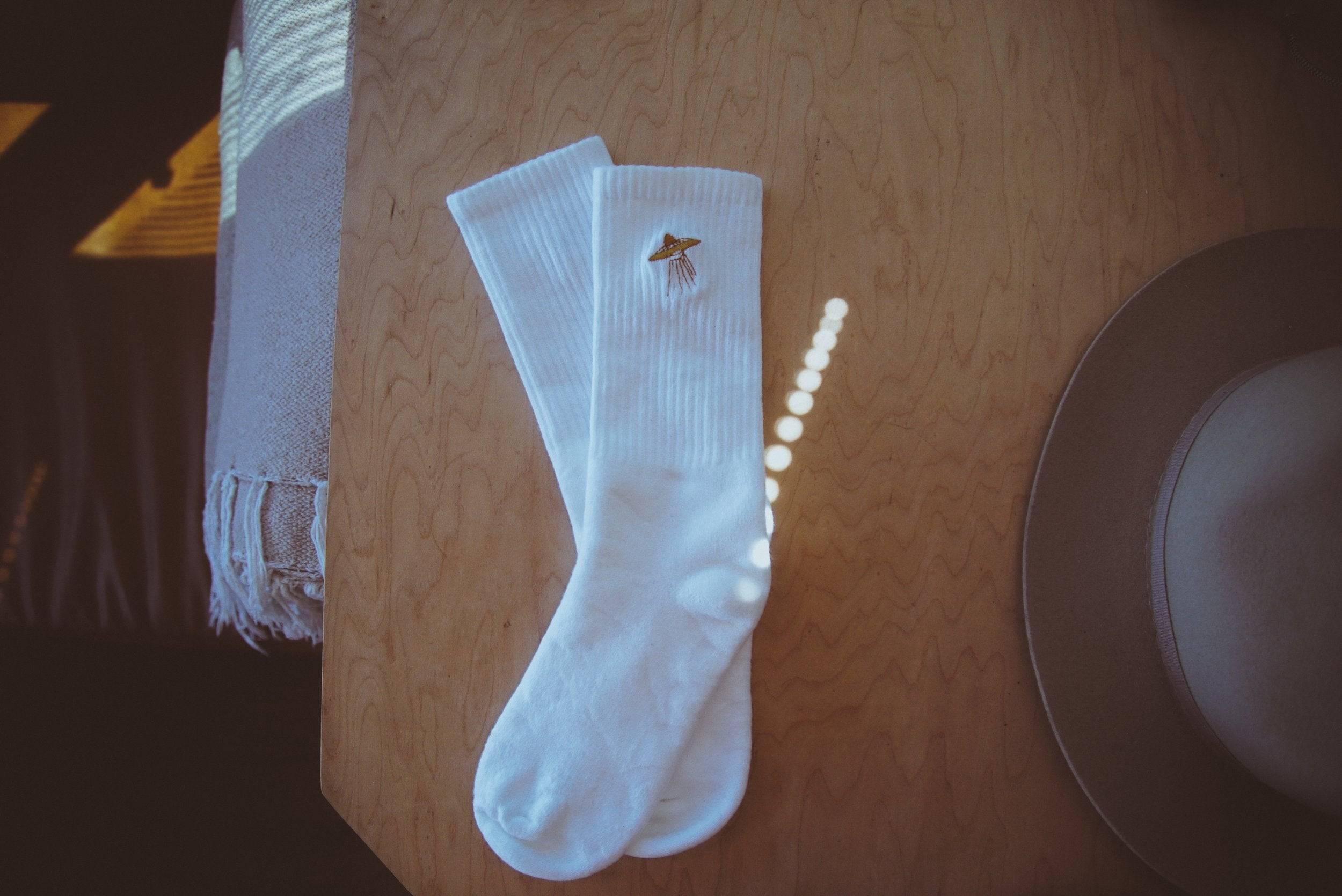 Pair of white ribbed socks with UFO embroidery on wooden surface next to beige felt hat
