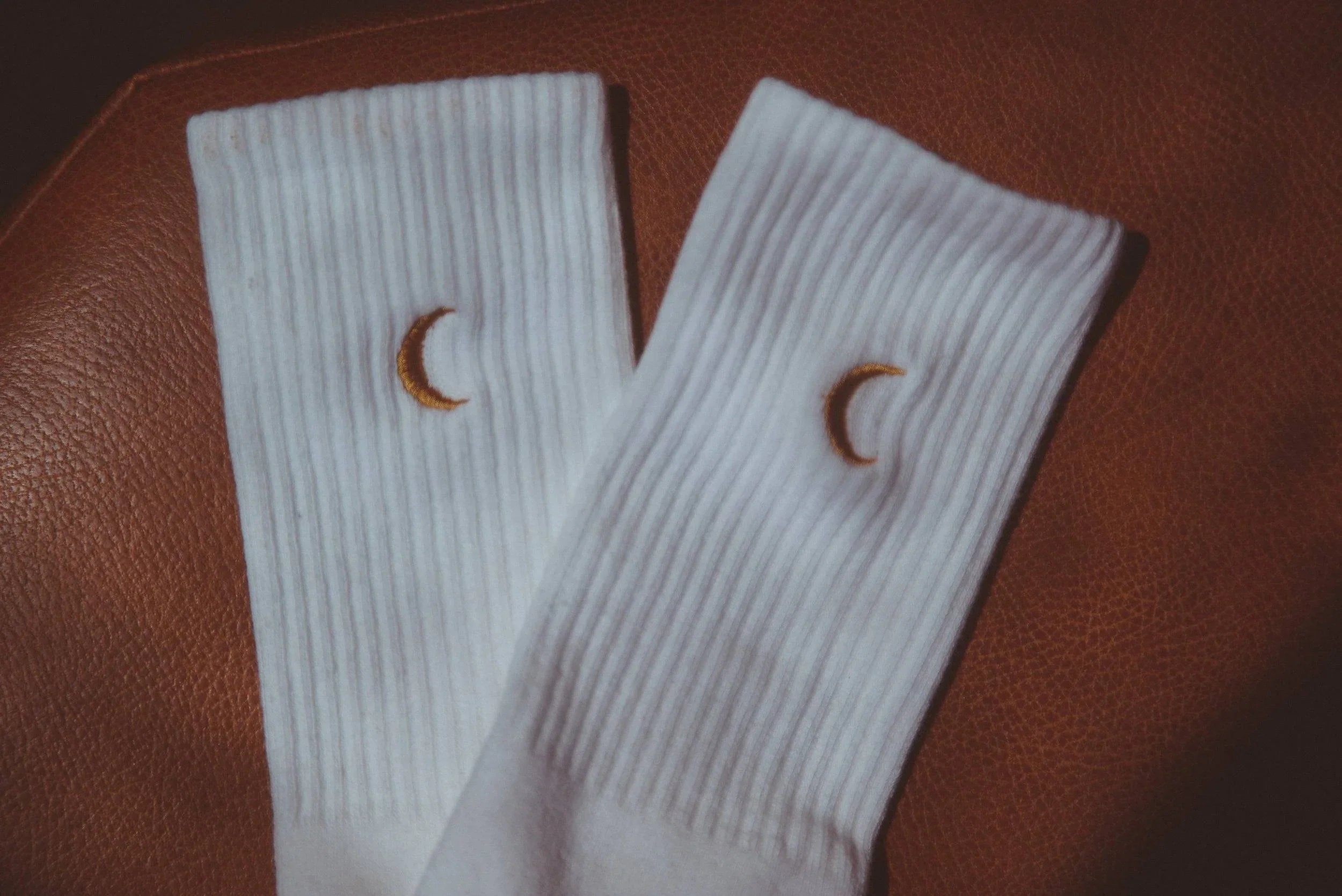 Pair of white ribbed socks with small embroidered crescent moons on brown leather surface