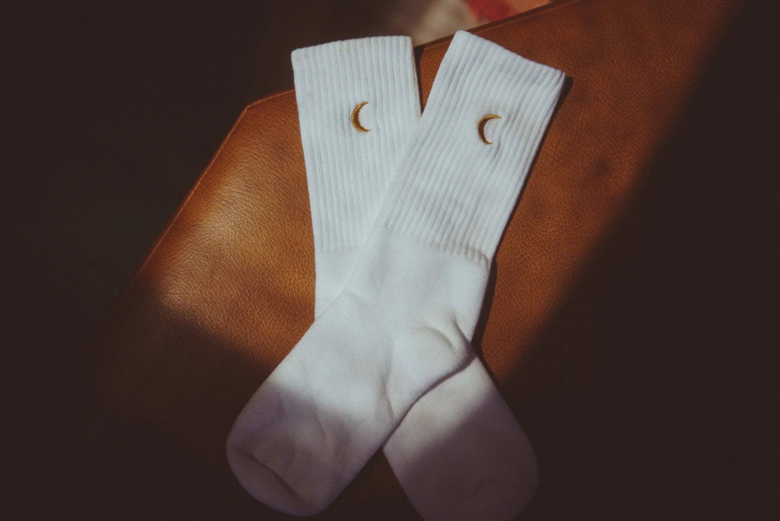 Pair of white ribbed crew socks with small golden crescent moon embroidery on brown leather surface