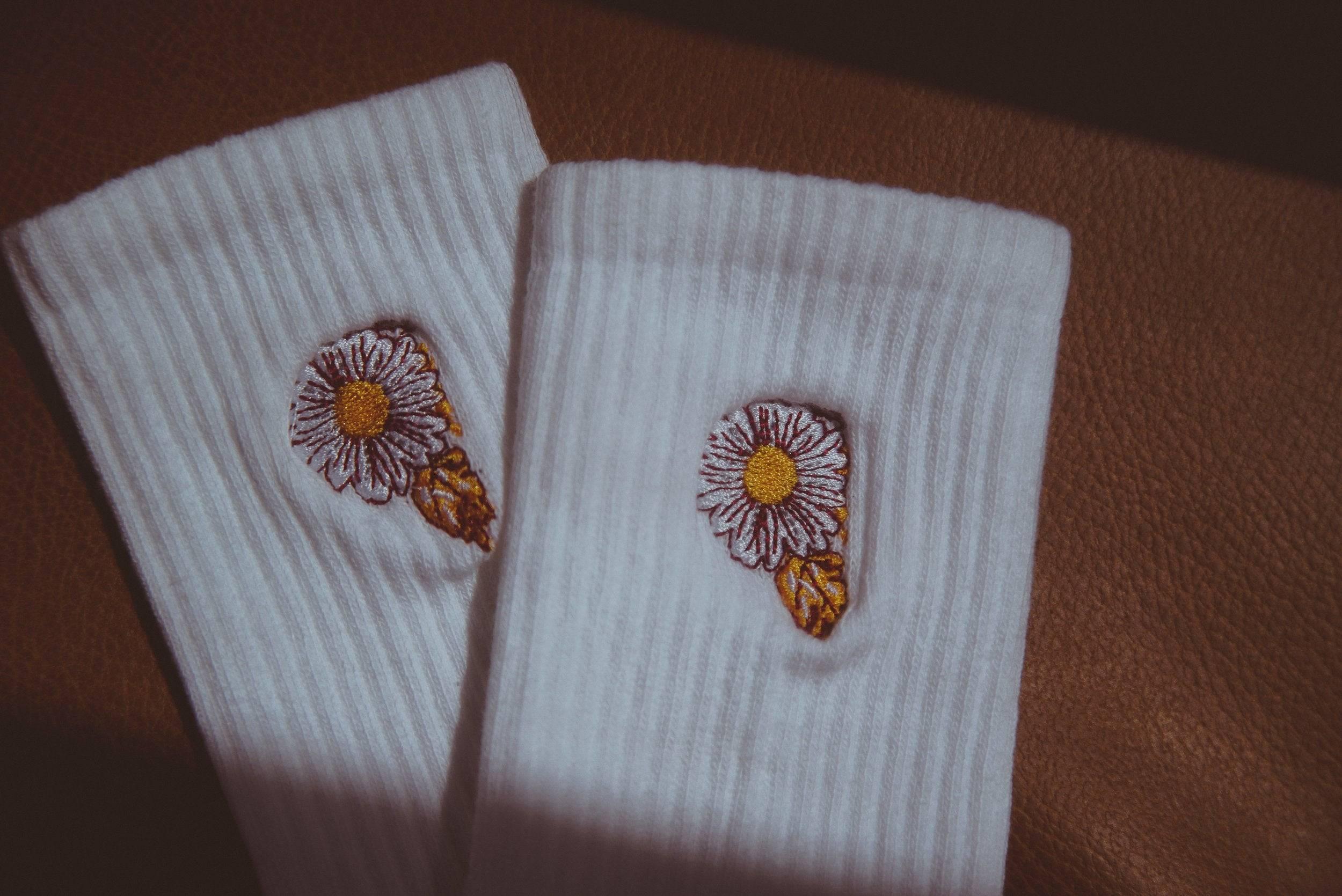 Pair of white ribbed socks with embroidered daisy flower and leaf design on brown surface