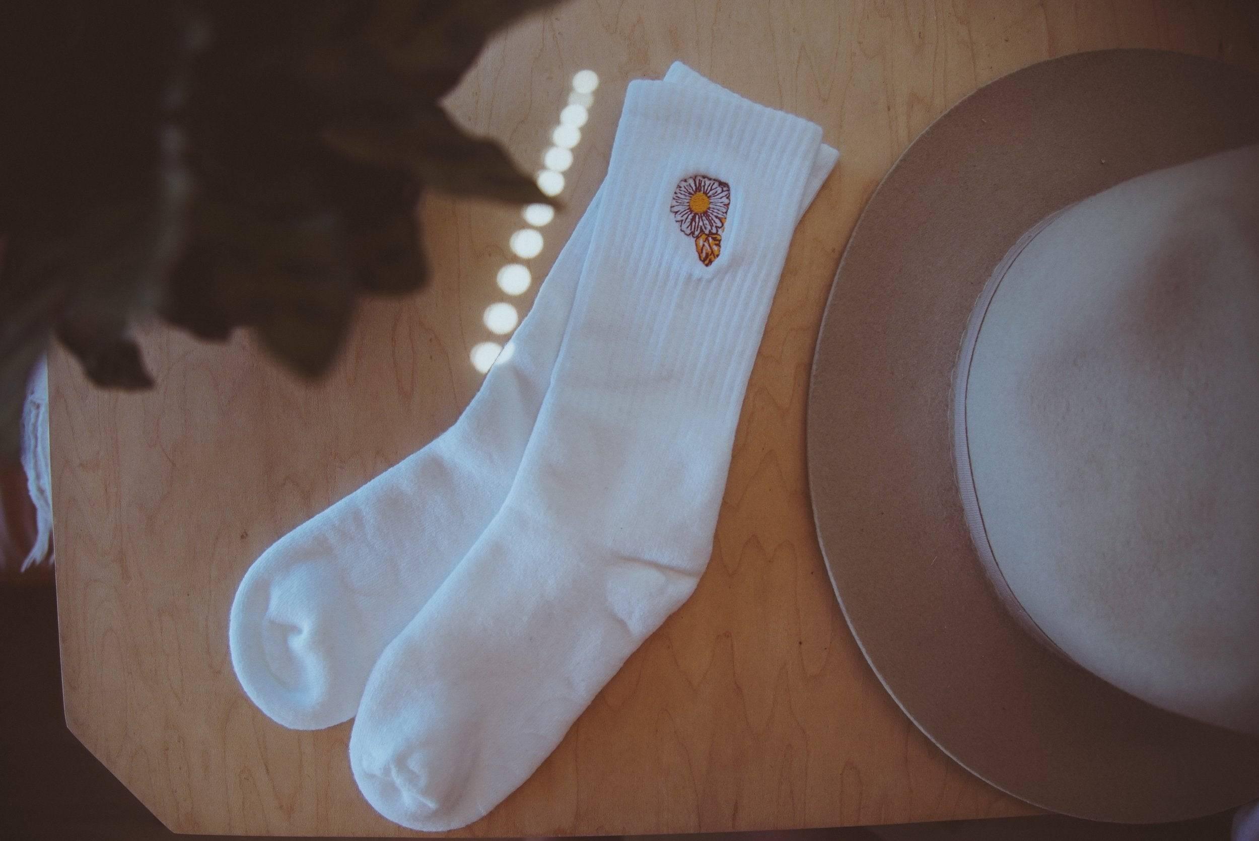 Pair of white ribbed socks with daisy embroidery on wooden table next to beige felt hat