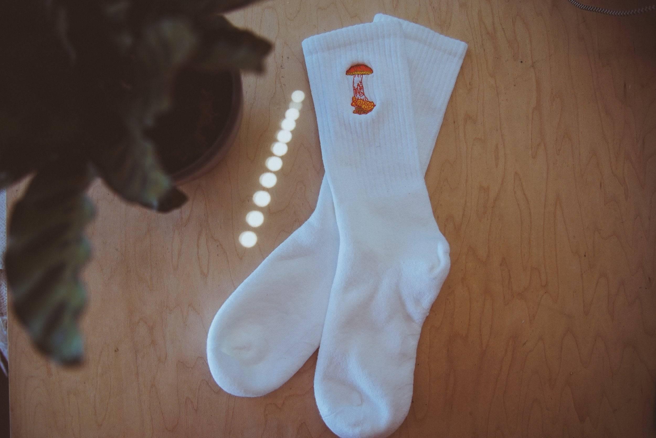 White socks with an embroidered orange mushroom design on wooden surface next to a potted plant