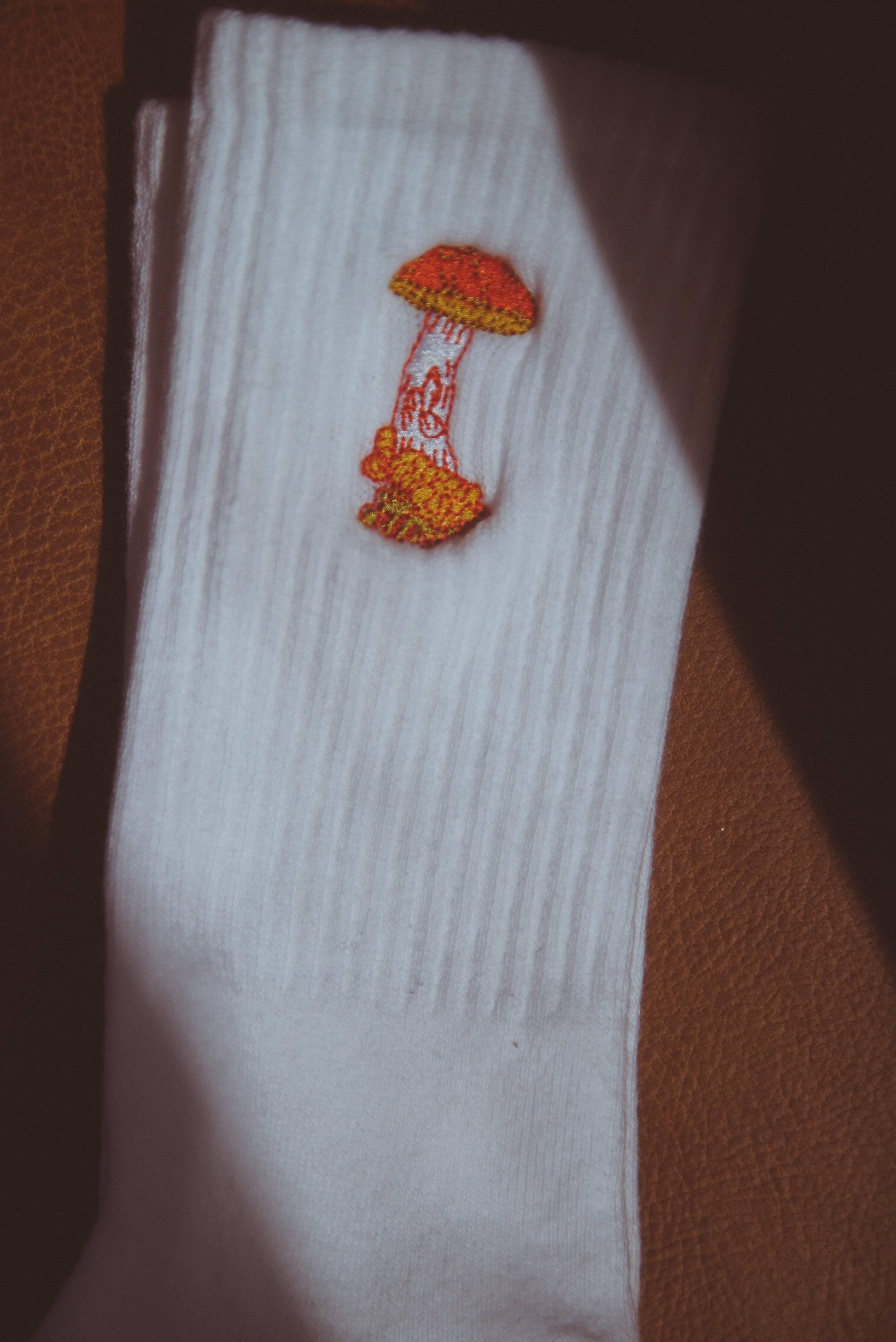 White ribbed sock with an embroidered orange and yellow mushroom design on brown leather background